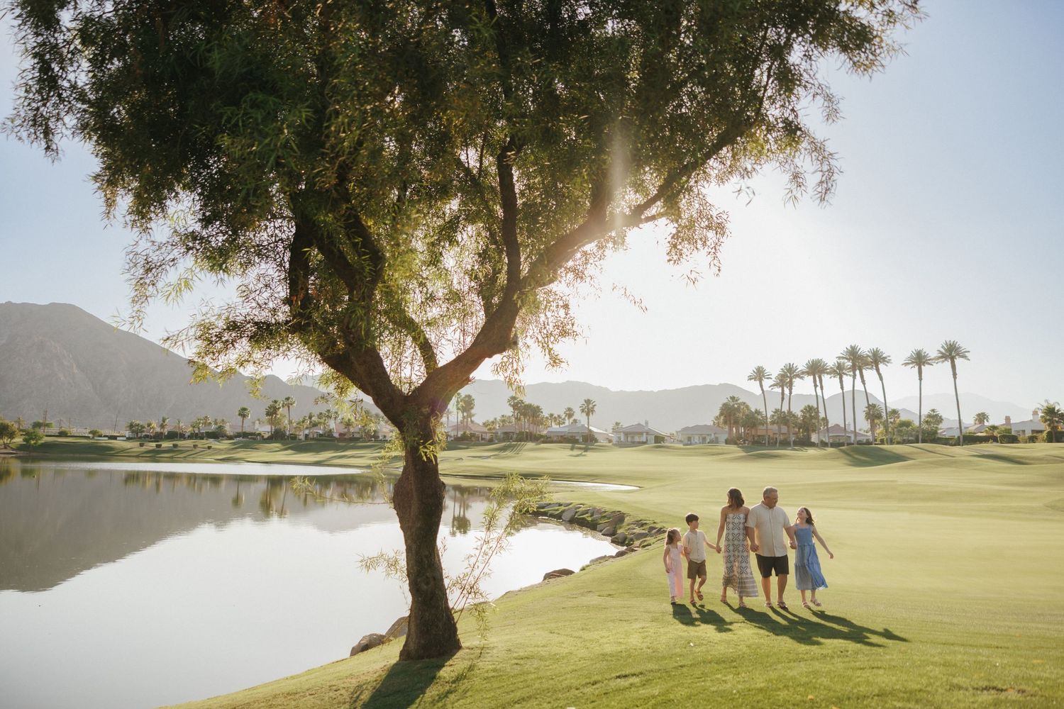 Family walking hand-in-hand on a lush golf course near a pond, with palm trees and mountains in the background.