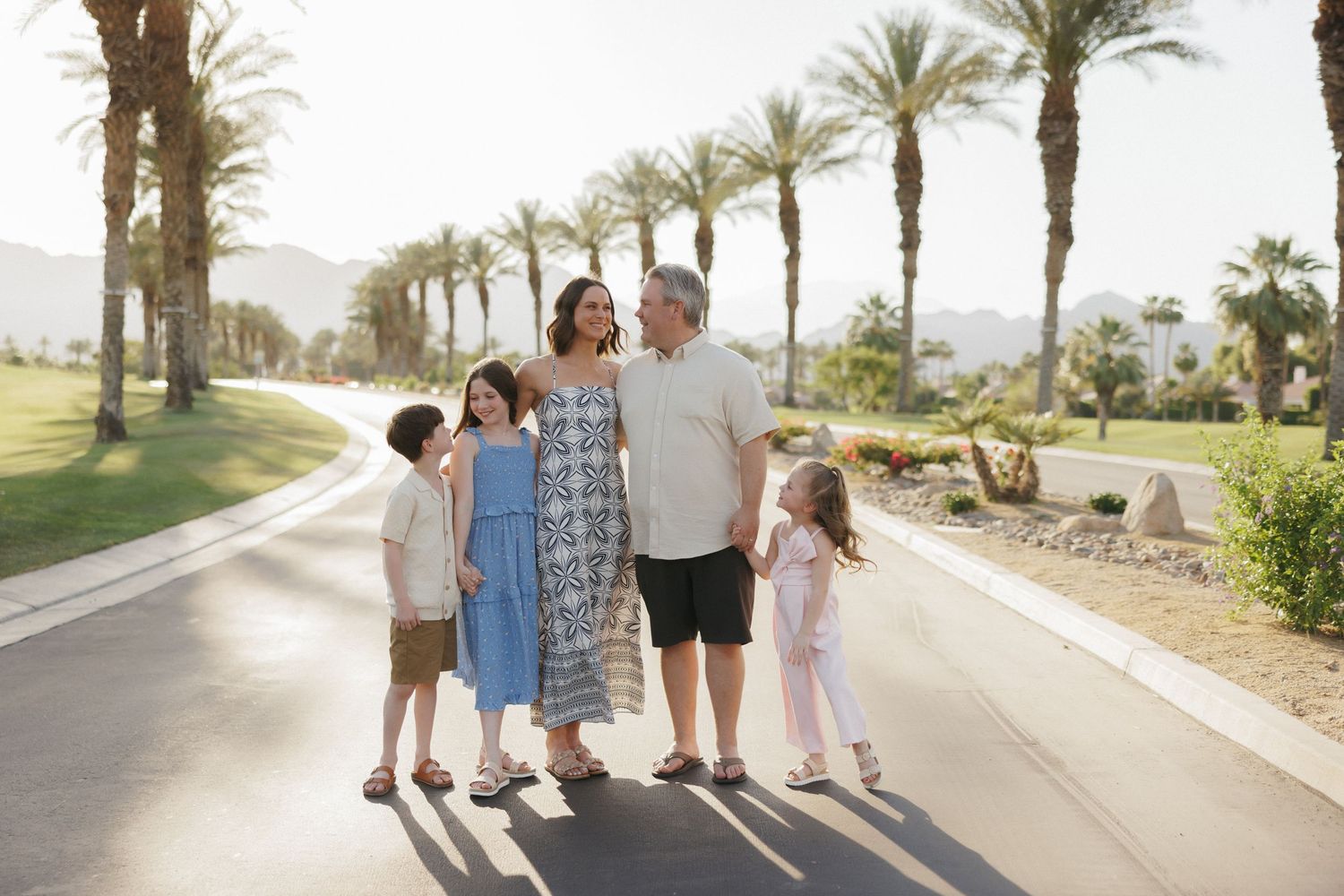 Family of five posing on a sunny palm-lined path with mountains in the background at a resort.