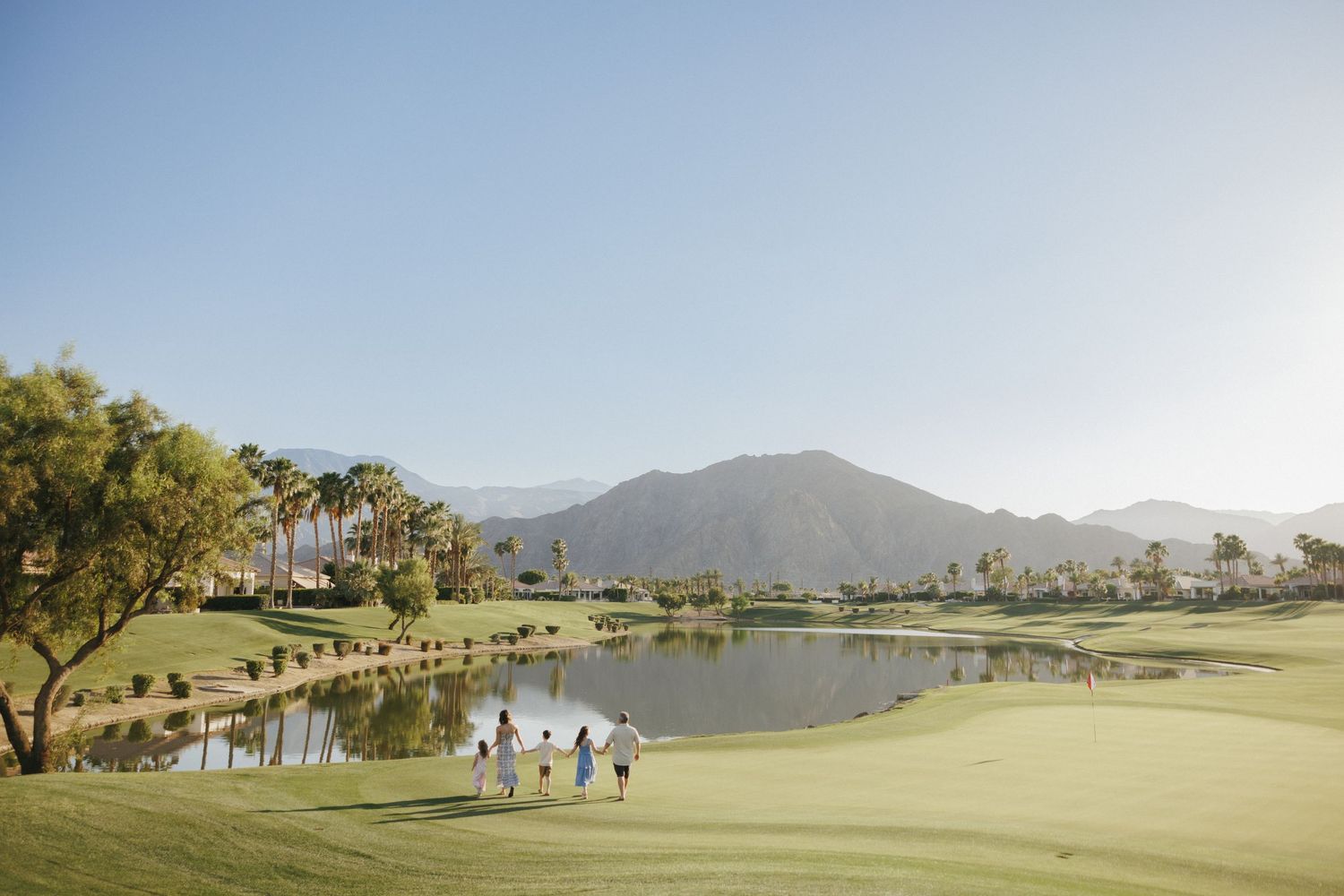 Families walking on a lush green golf course with a scenic lake and mountain backdrop under a clear blue sky.