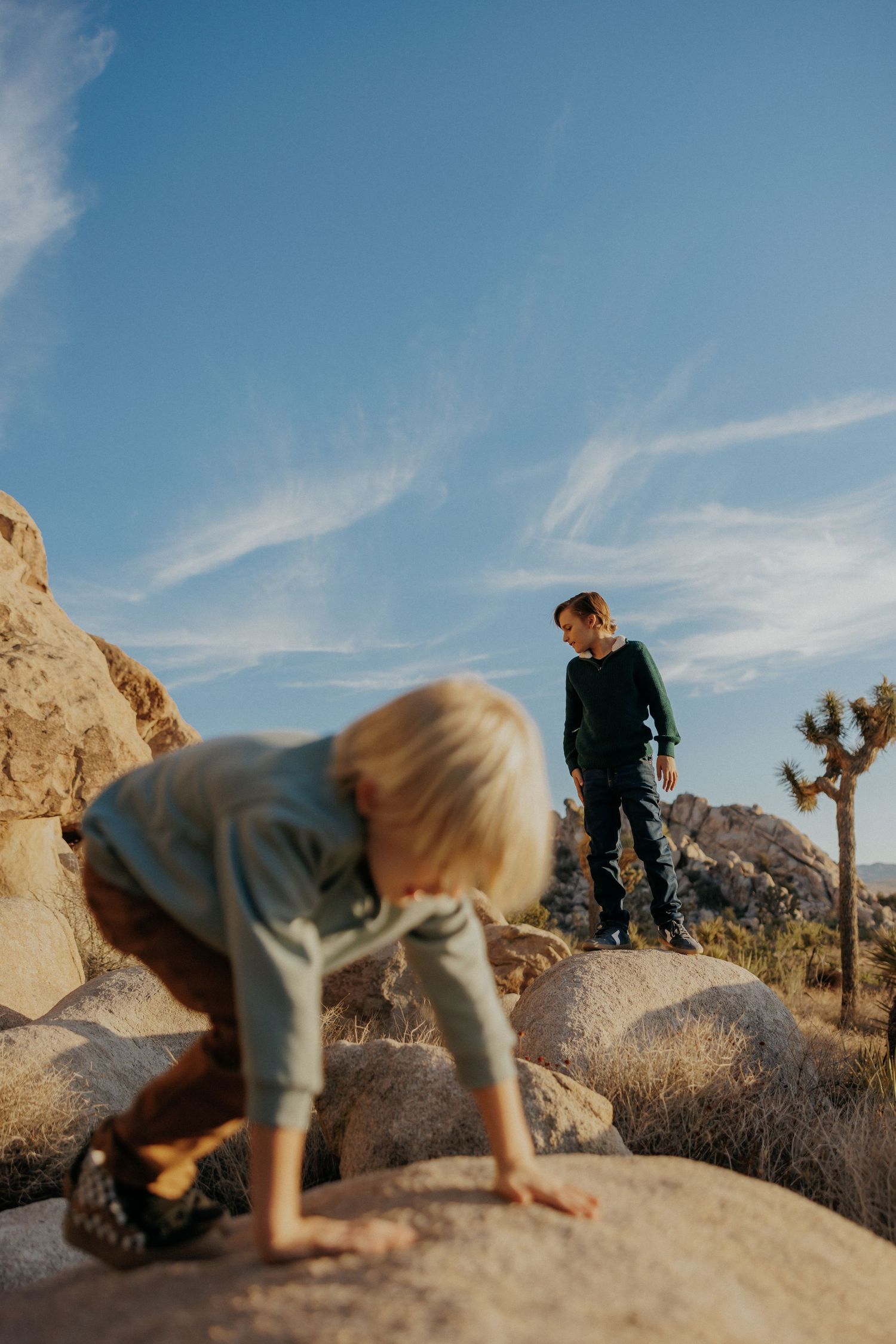 Two children climbing rocks in Joshua Tree National Park under a blue sky with wispy clouds.