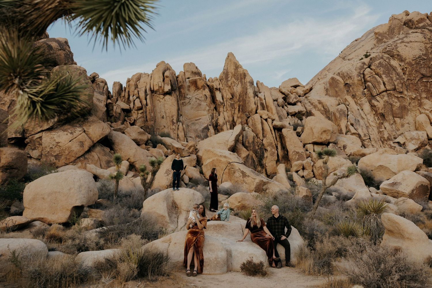 Group of people gathered among large boulders and Joshua trees in a desert landscape under a cloudy sky.