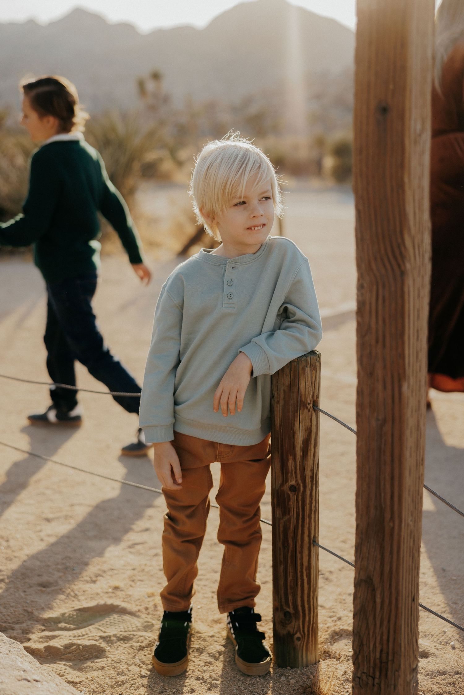 Blonde boy in light blue henley and brown pants leans against wooden post in desert landscape.