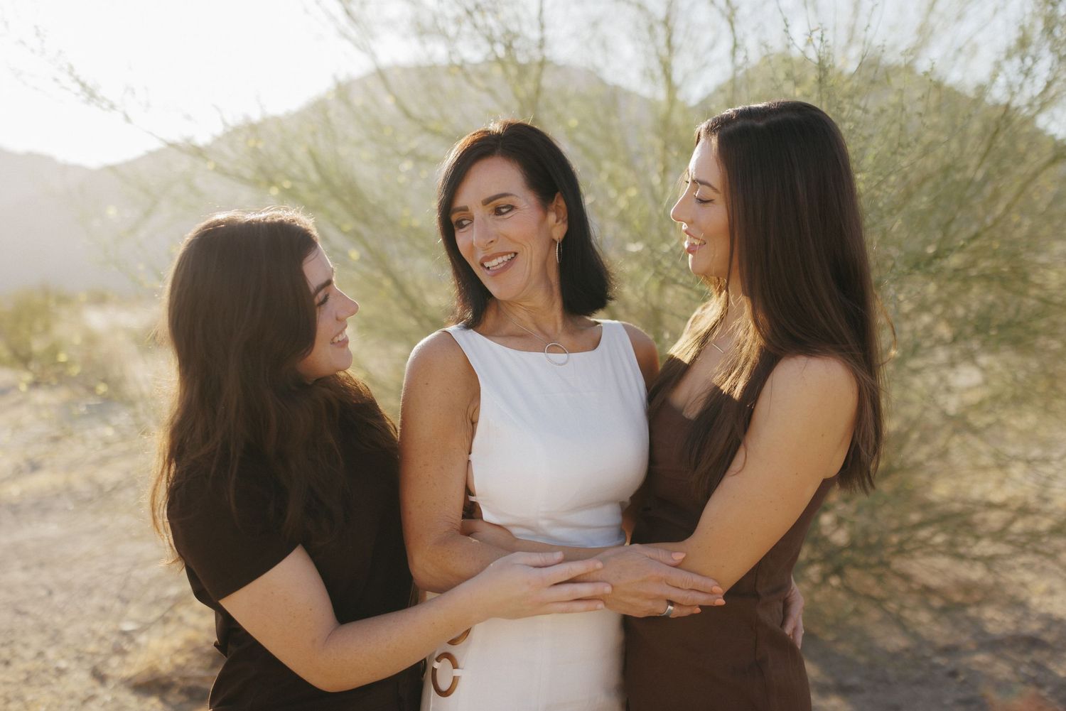 Three women smiling and embracing outdoors in a desert landscape with warm golden sunlight in the background.
