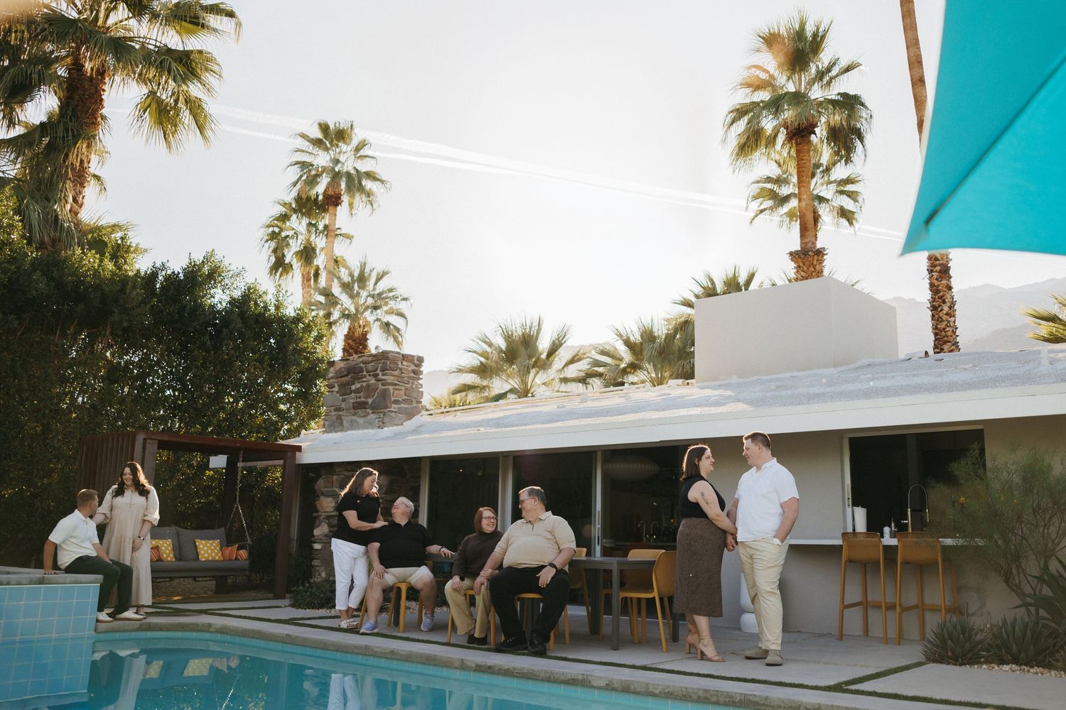 Group of people socializing by a pool at a mid-century modern home surrounded by palm trees.