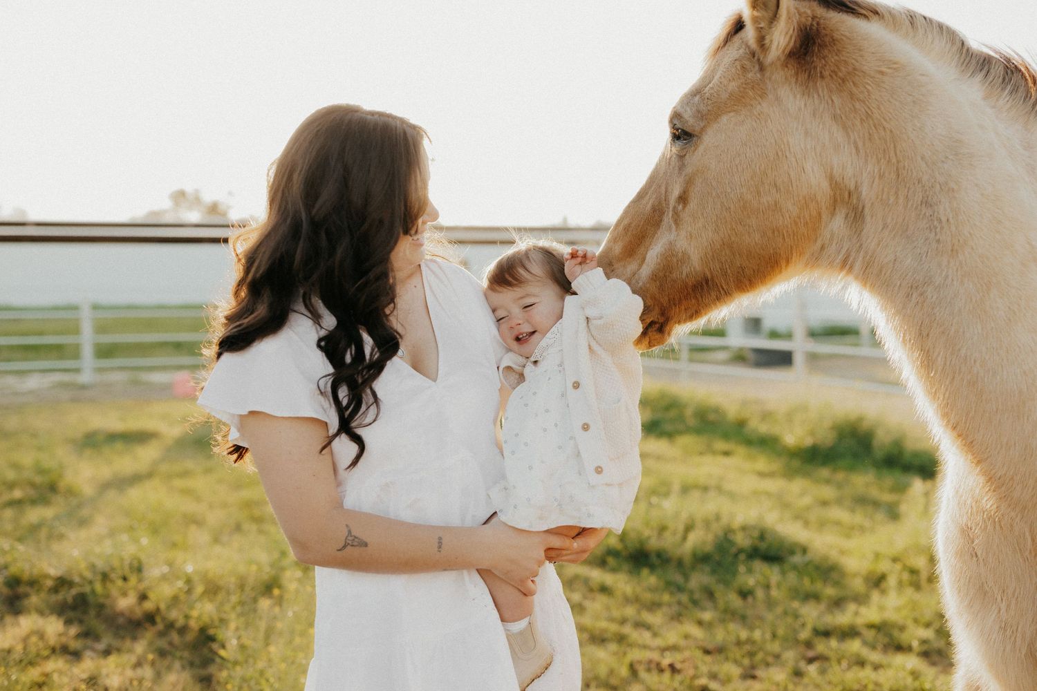 A woman in a white dress holds a baby while a palomino horse gently nuzzles the child outdoors.