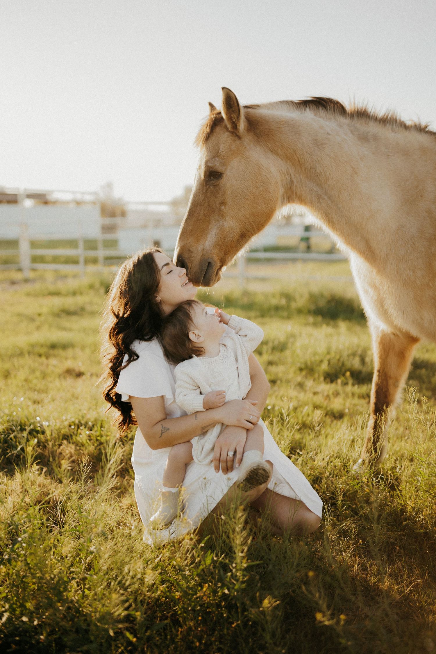 A mother sits in a green field holding her child while a palomino horse nuzzles her face near a white fence.