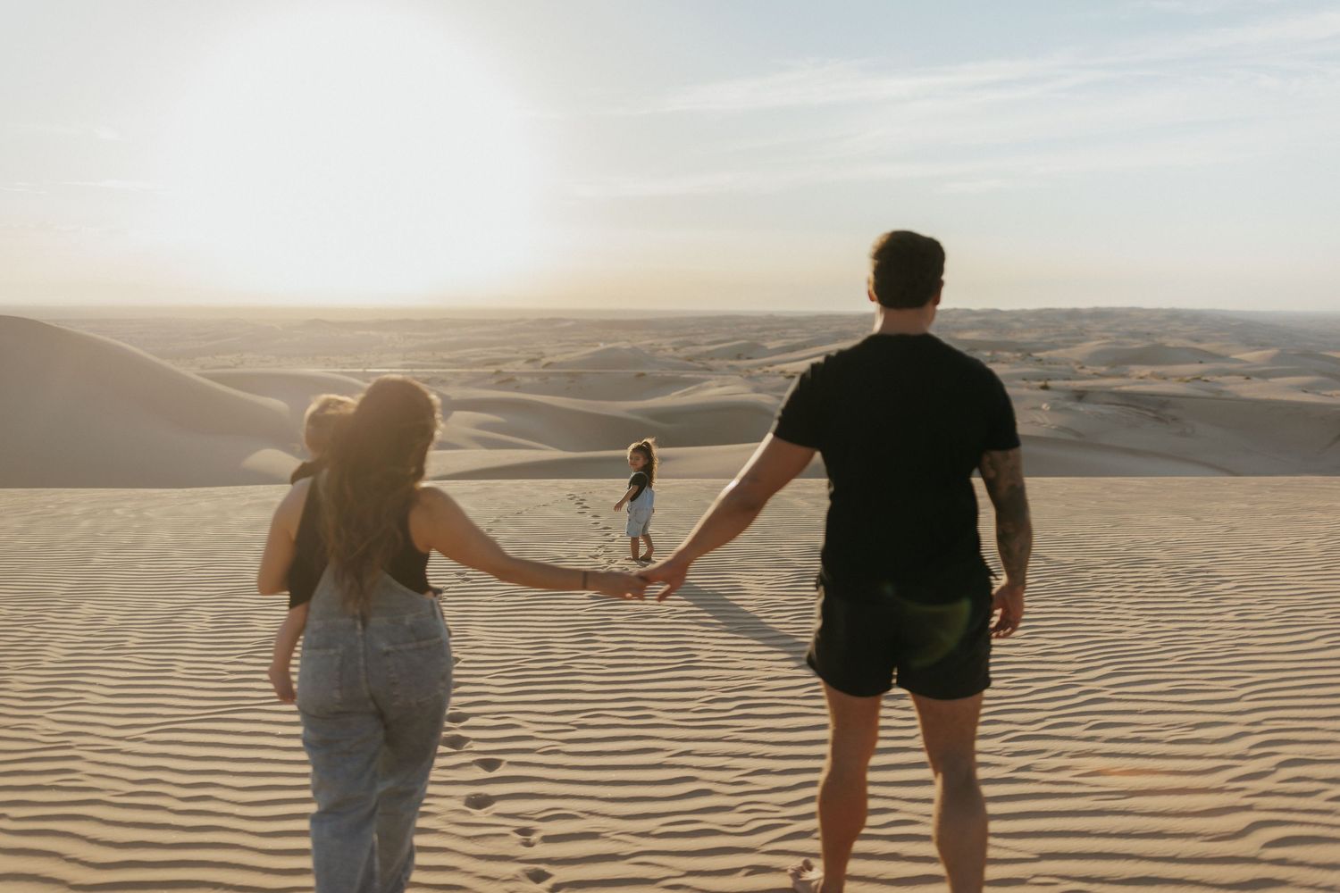 Couple holding hands on sandy desert dunes at sunset with a child standing ahead in the golden landscape.
