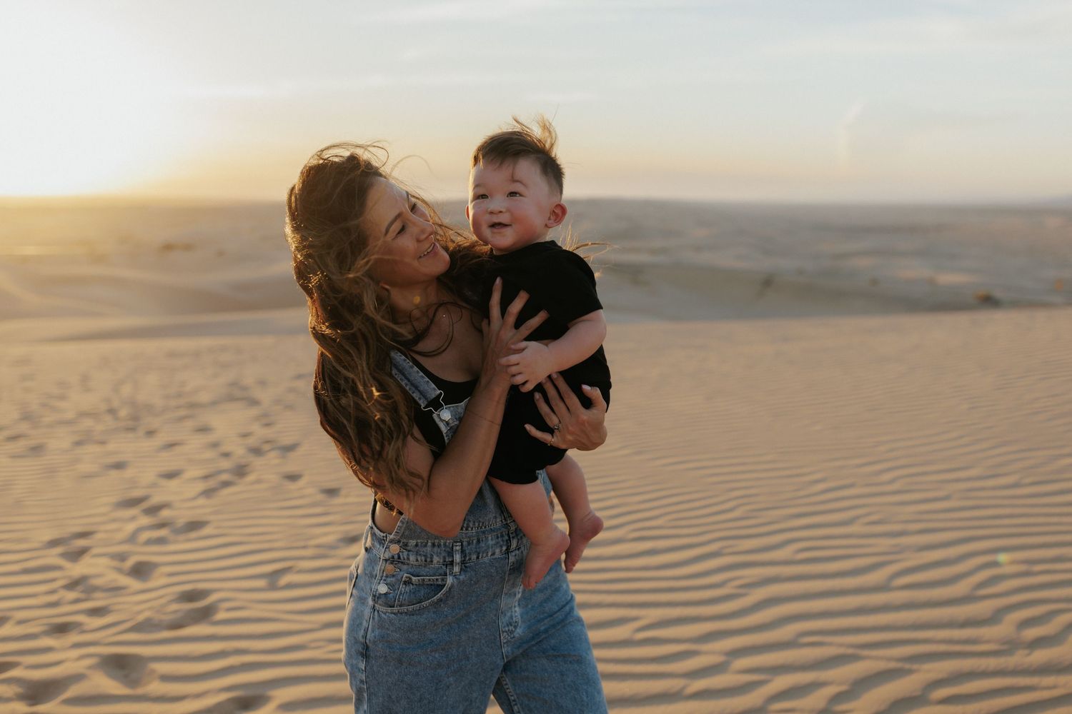 Woman holding baby on sandy desert dunes at golden hour sunset, wearing jeans and black shirt.