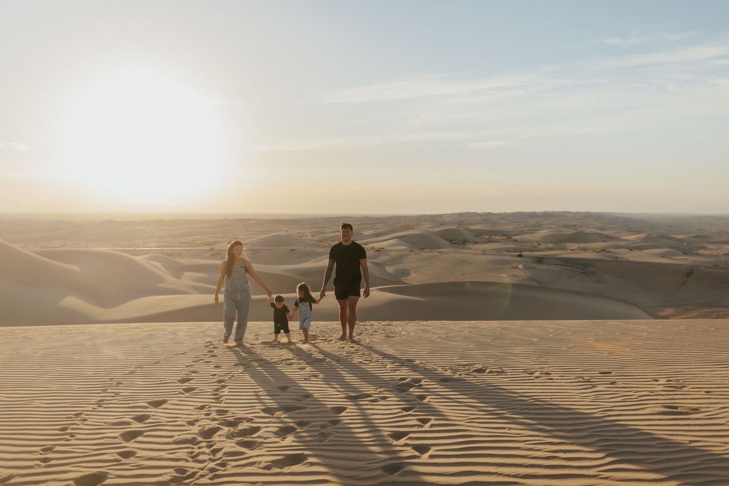 Family of three holding hands walking on golden sand dunes at sunset in a vast desert landscape.