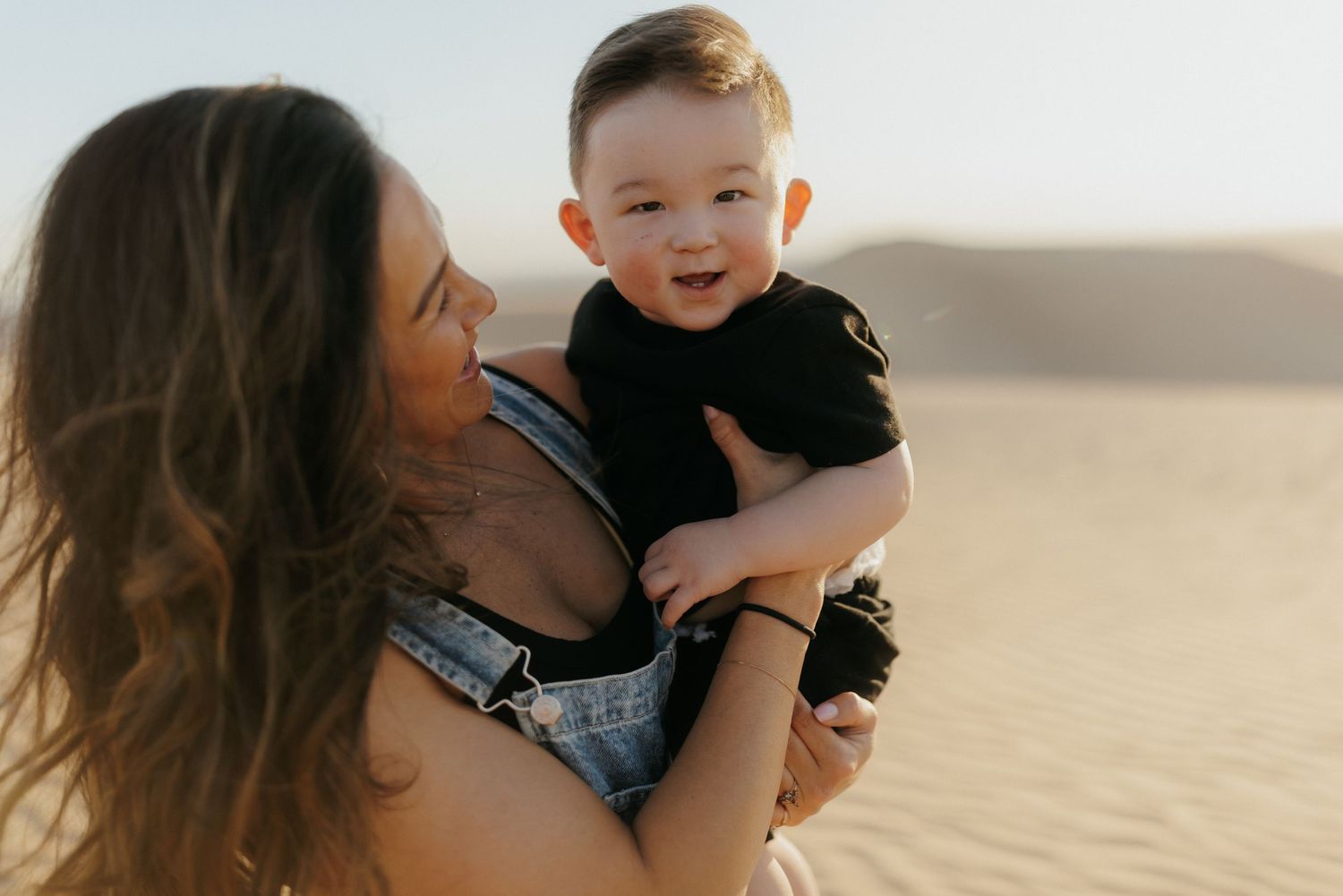 A mother holds her smiling baby boy on sandy desert dunes during a golden hour outdoor photoshoot.