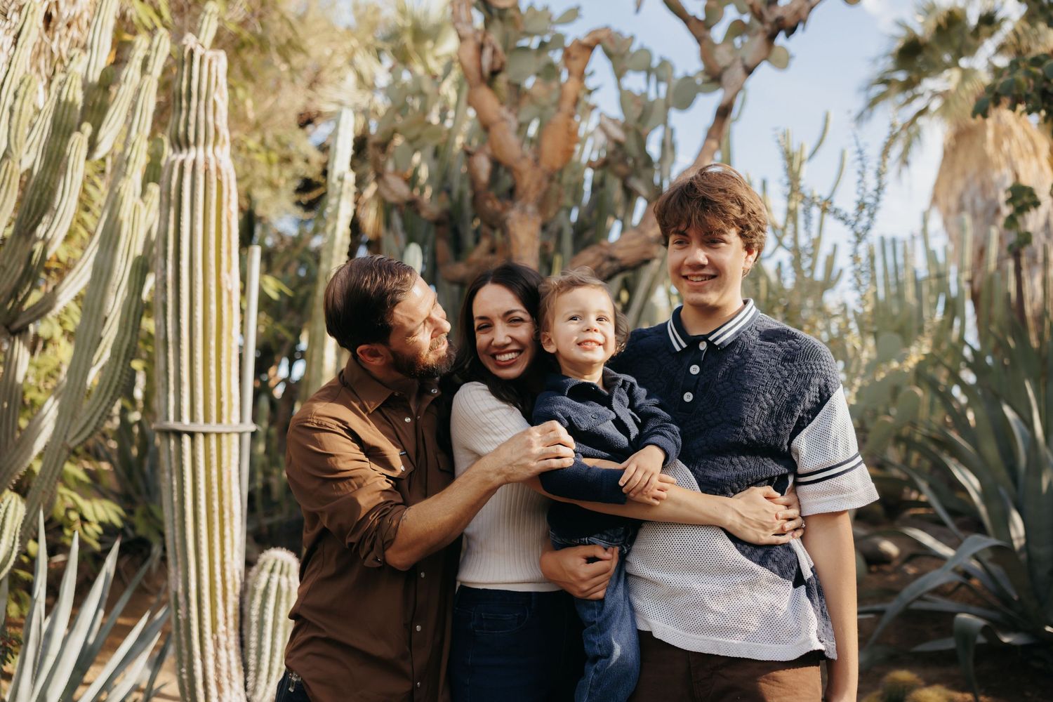 Family of four smiling outdoors among tall cacti and palm trees on a sunny day.