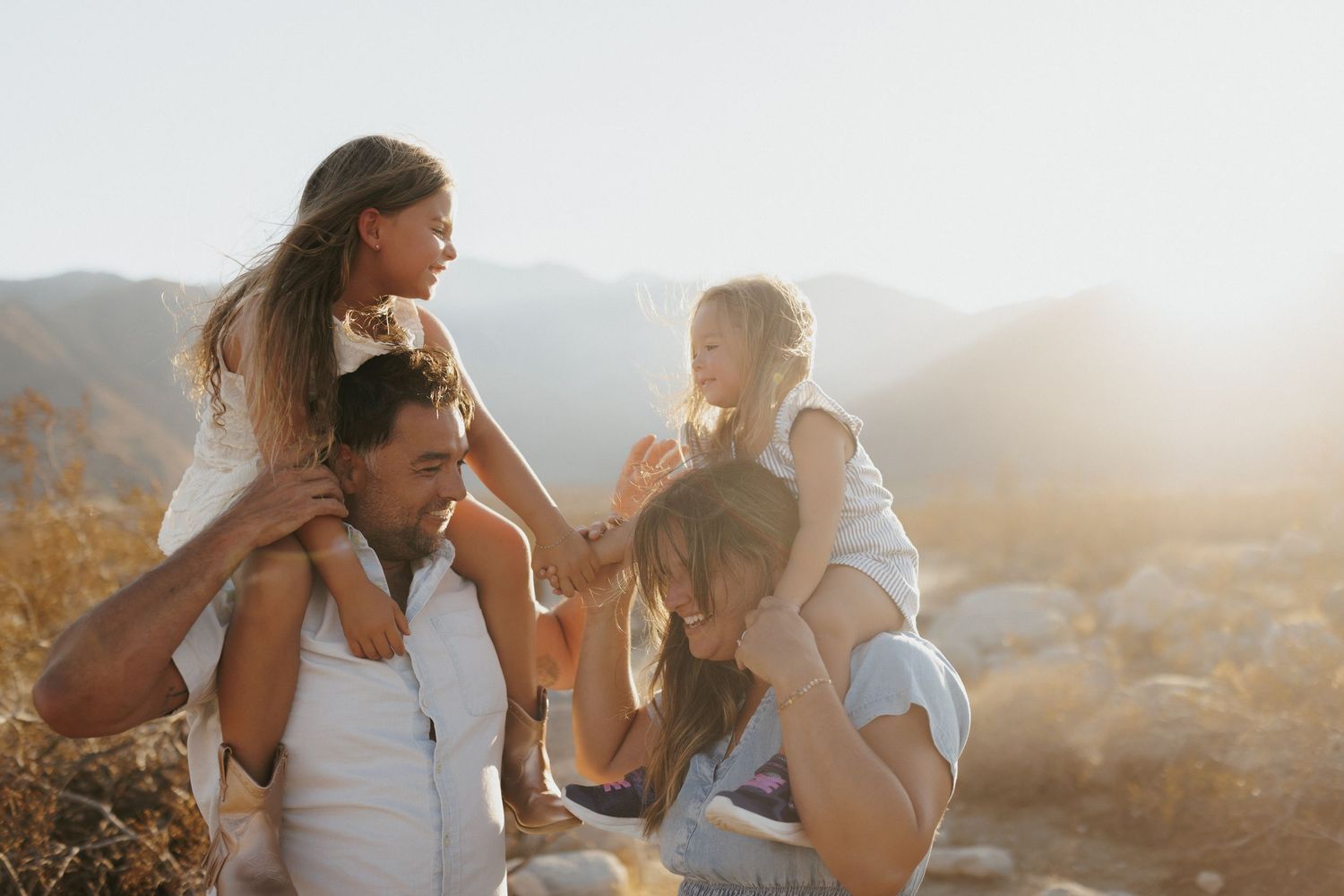Family of four enjoying a sunny mountain outing, with children on parents shoulders, laughing together outdoors.