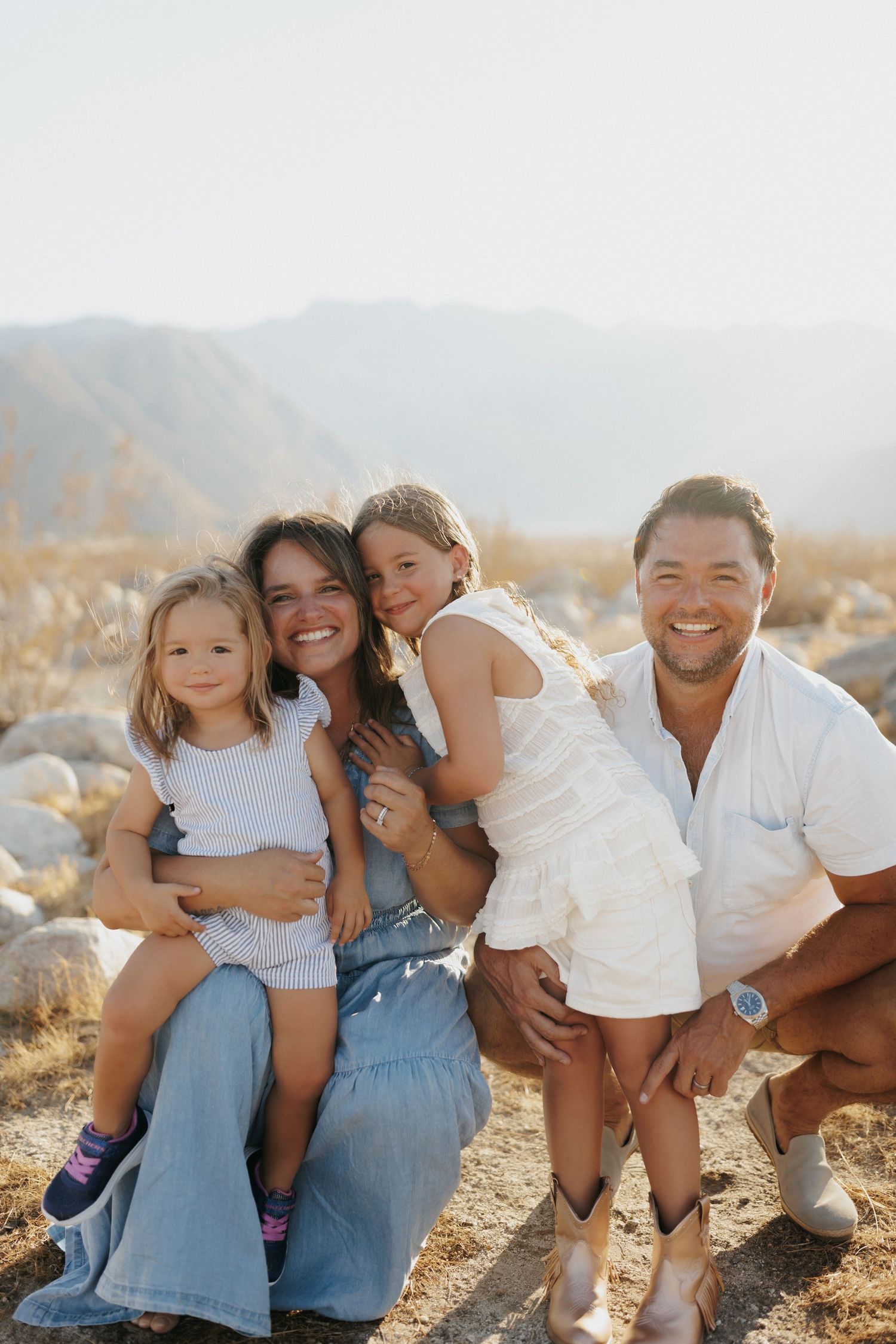 Happy family of four posing outdoors with mountain backdrop on sunny day, parents holding young children.