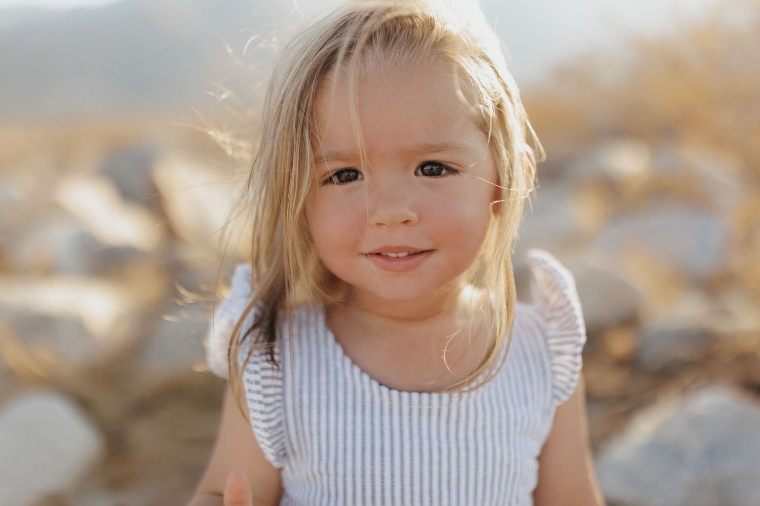 Smiling toddler girl with blonde hair posing outdoors among rocks in warm sunlight.