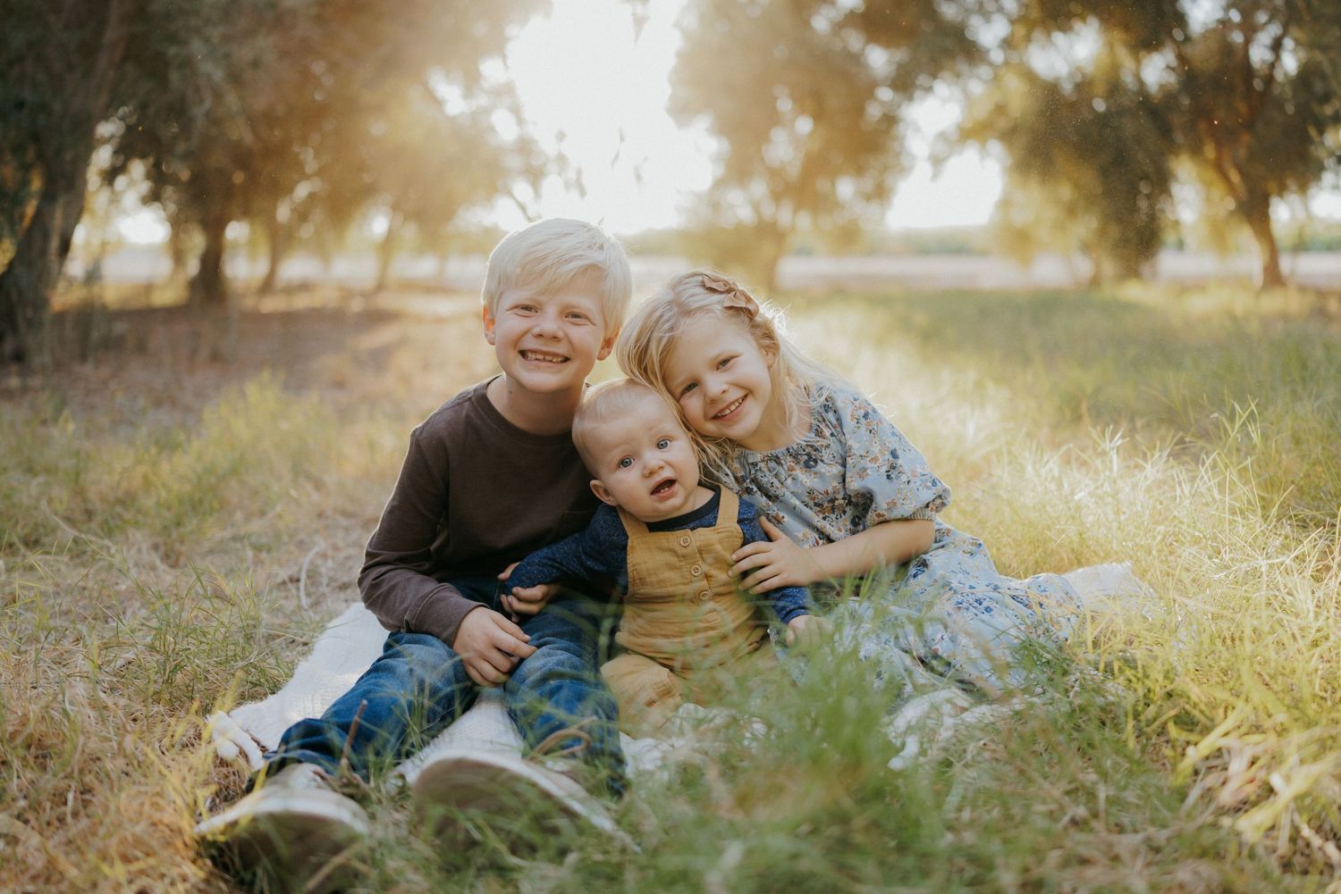 Three young siblings sit together in a sunlit field, smiling warmly, surrounded by golden grass and trees.