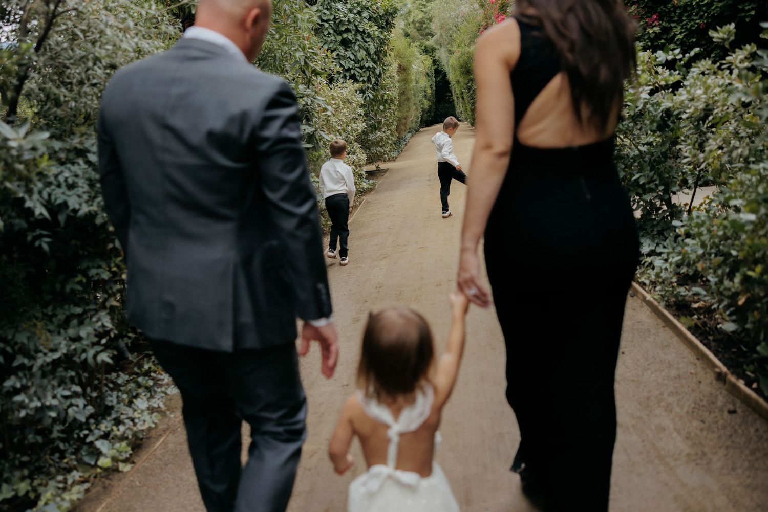 Family walking hand-in-hand with toddler along garden path surrounded by green hedges.