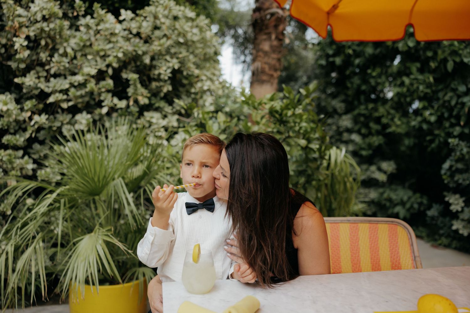 A mother kisses her young boy in a bow tie at an outdoor table surrounded by lush greenery.
