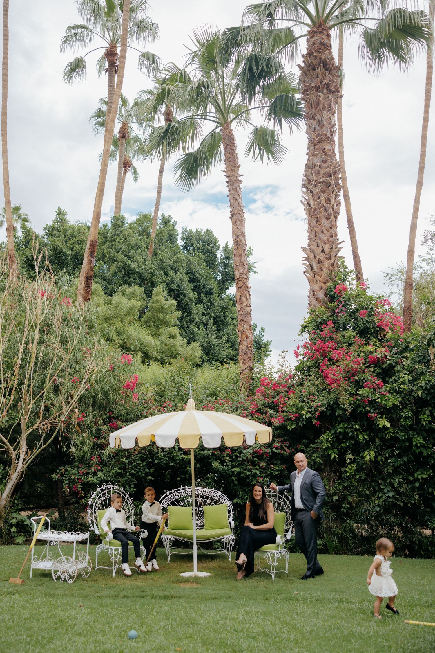 Family gathered under striped umbrella on lush garden lawn with tall palm trees and pink flowering bushes.