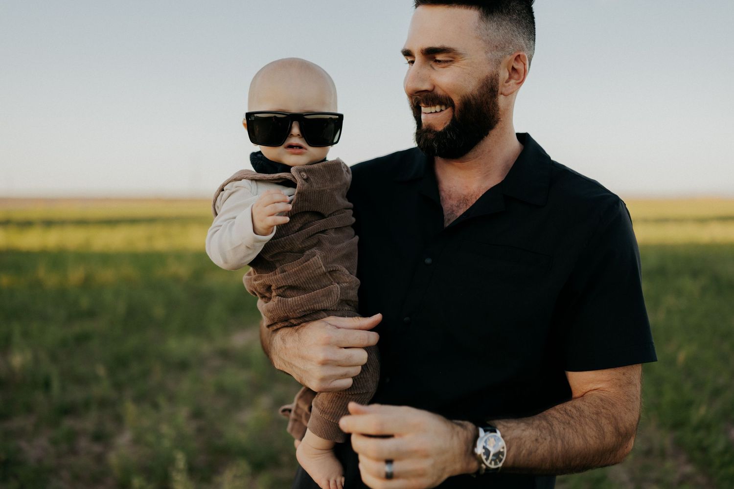 Smiling bearded man holding a baby wearing sunglasses in a green field on a sunny day.