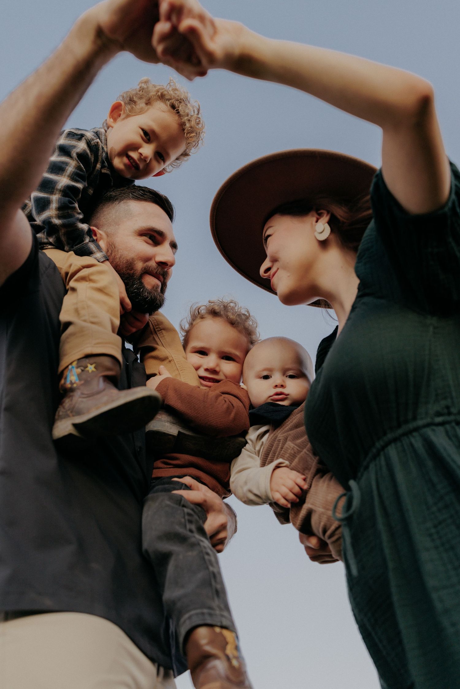 Happy family with three young children outdoors, parents smiling and holding kids against a blue sky.