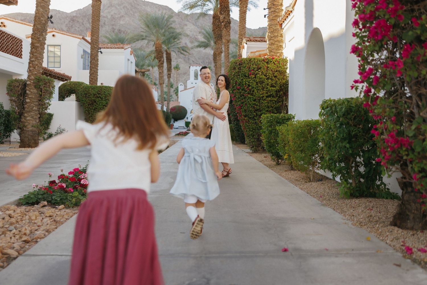 Family reunion outdoors near white stucco buildings with palm trees, children running toward parents on a sunny day.