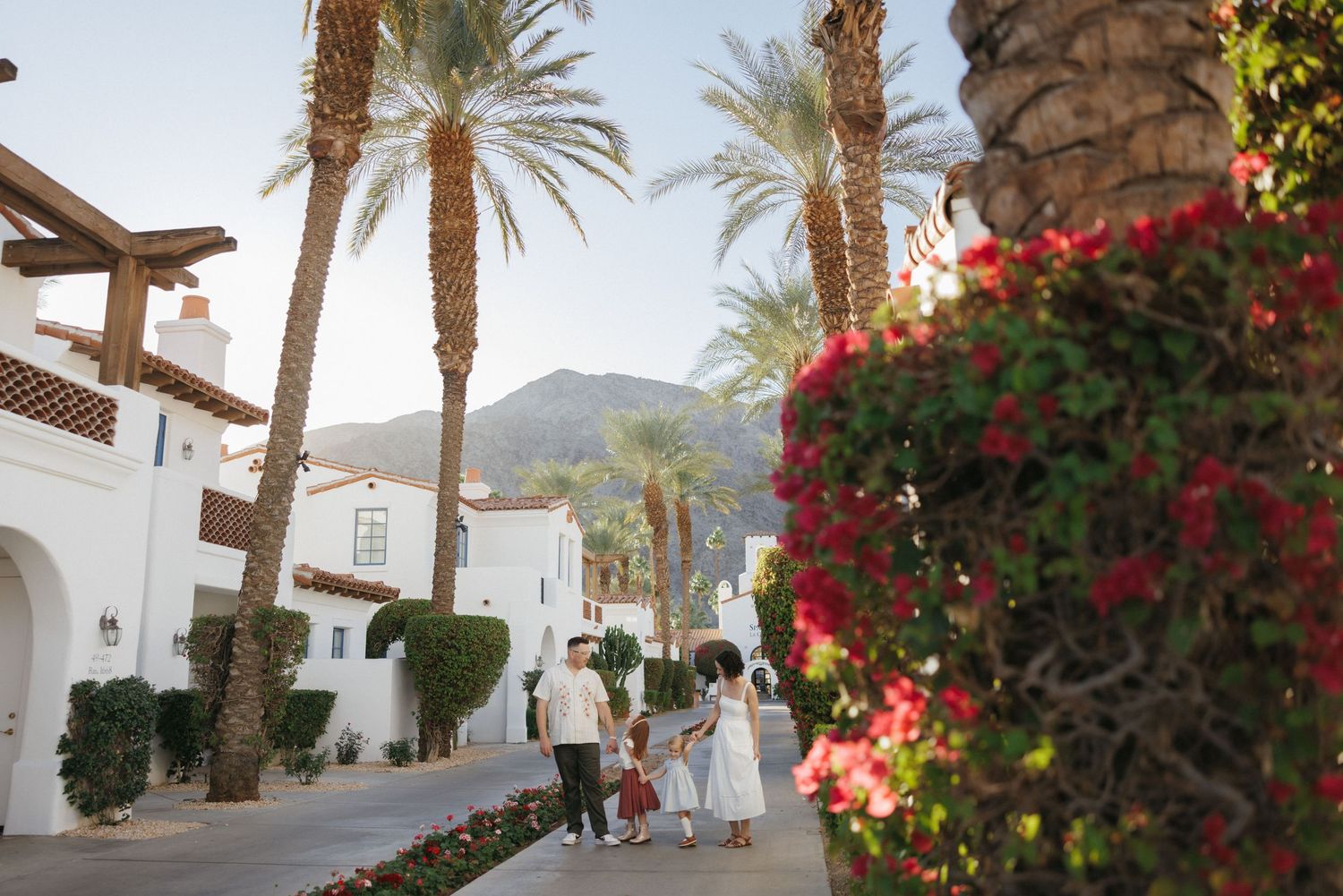 Family strolling past white Spanish-style villas lined with tall palm trees and red bougainvillea, mountains in background.
