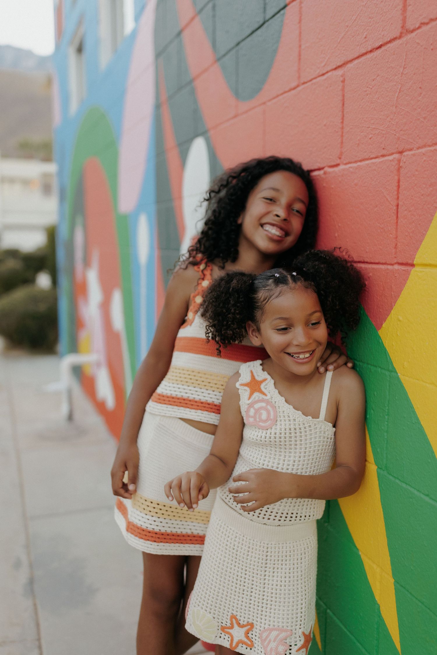 Two young girls in crochet dresses smiling and posing together against a colorful painted mural wall.