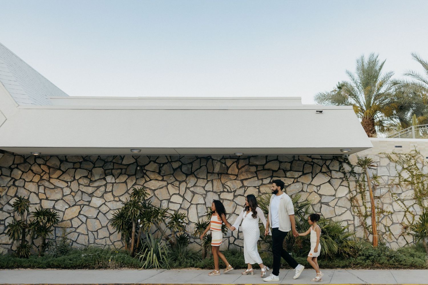 Family of four walking past a stone wall with palm trees and a modern white building in a sunny outdoor setting.