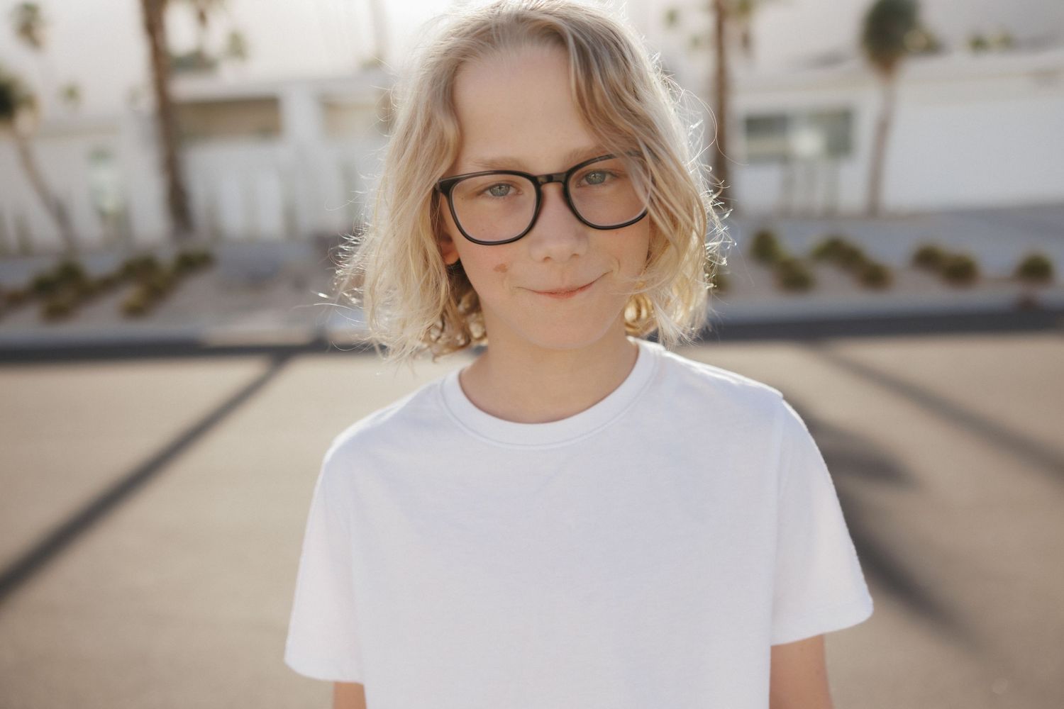 Young person with blonde bob haircut wearing glasses and white t-shirt, standing outdoors in sunny weather.