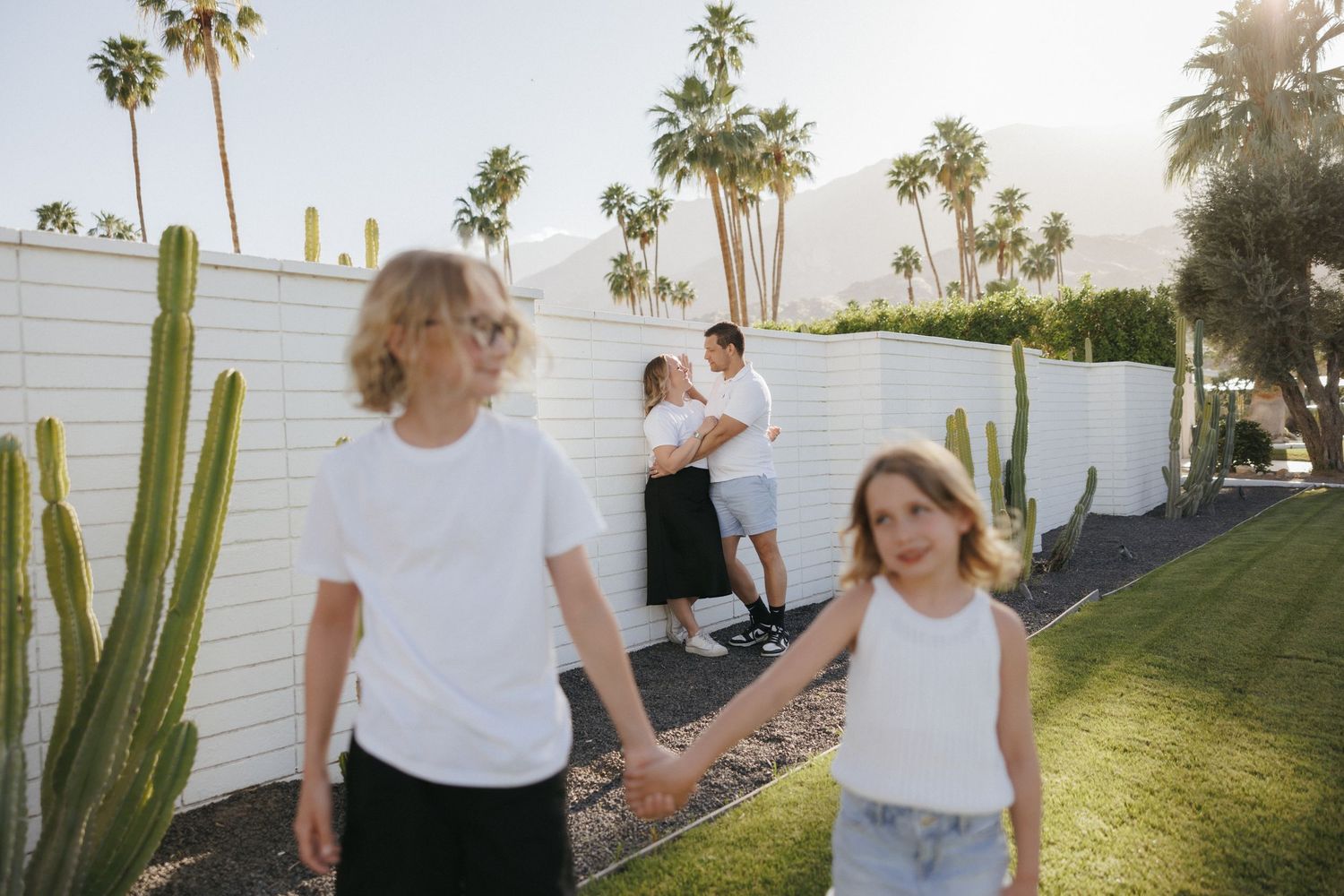 Two children hold hands in a sunny Palm Springs backyard with cacti, white walls, and palm trees while adults embrace behind them.