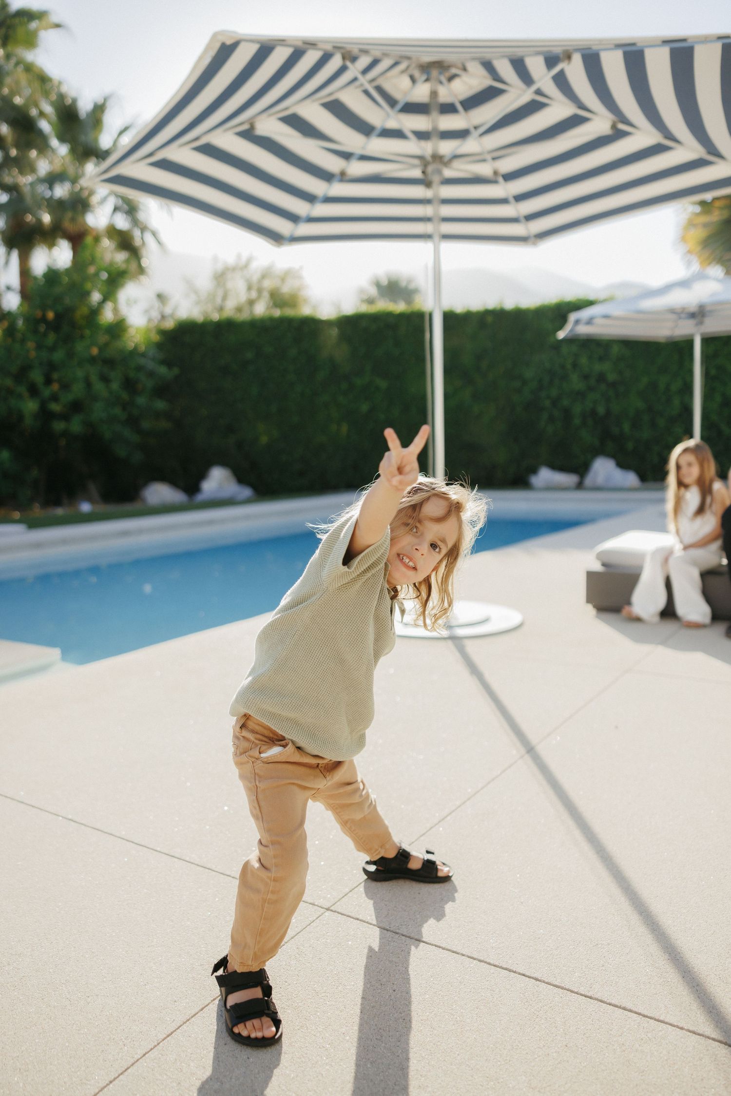 A young child making a peace sign by a sunny poolside with a striped umbrella and hedges in the background.