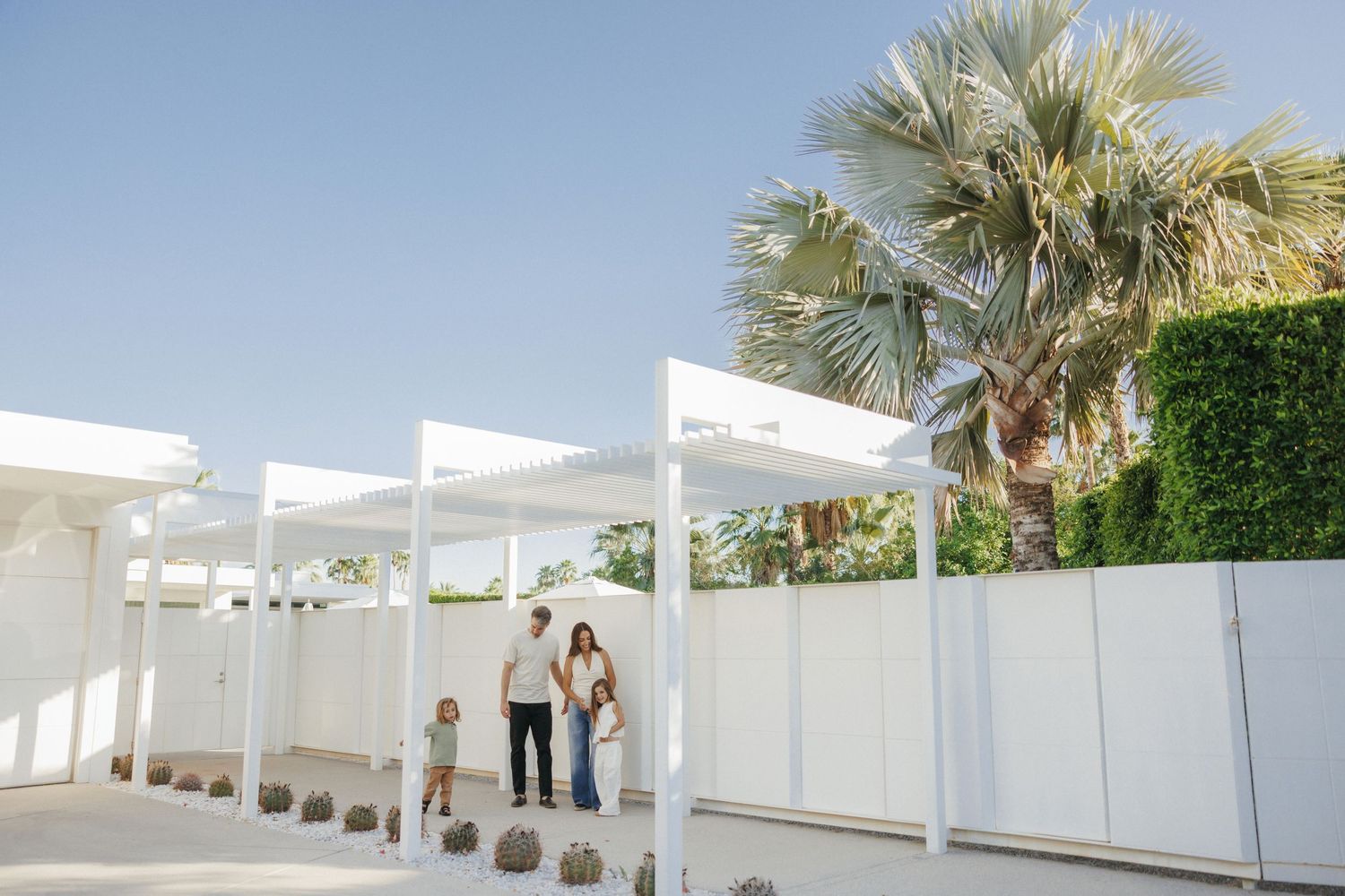 Family of three posing under white pergola with palm trees and lush greenery at a bright outdoor venue.