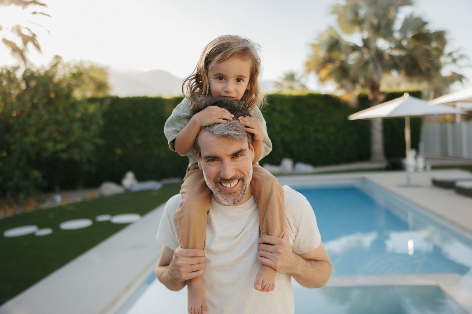 A smiling father carries his young daughter on his shoulders by a backyard pool surrounded by lush hedges.