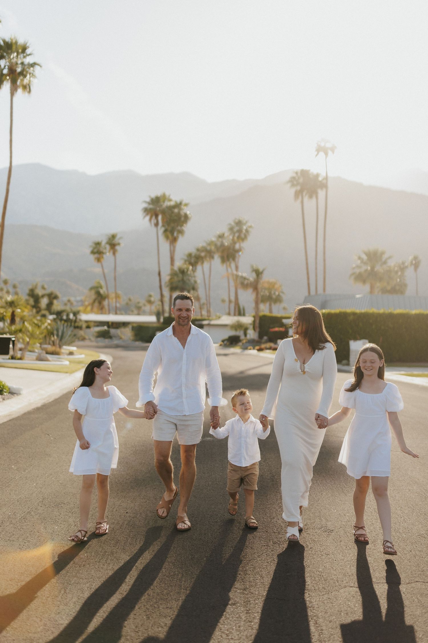 Family of five in white outfits walking hand-in-hand on a sunny Palm Springs street with mountains and palm trees.