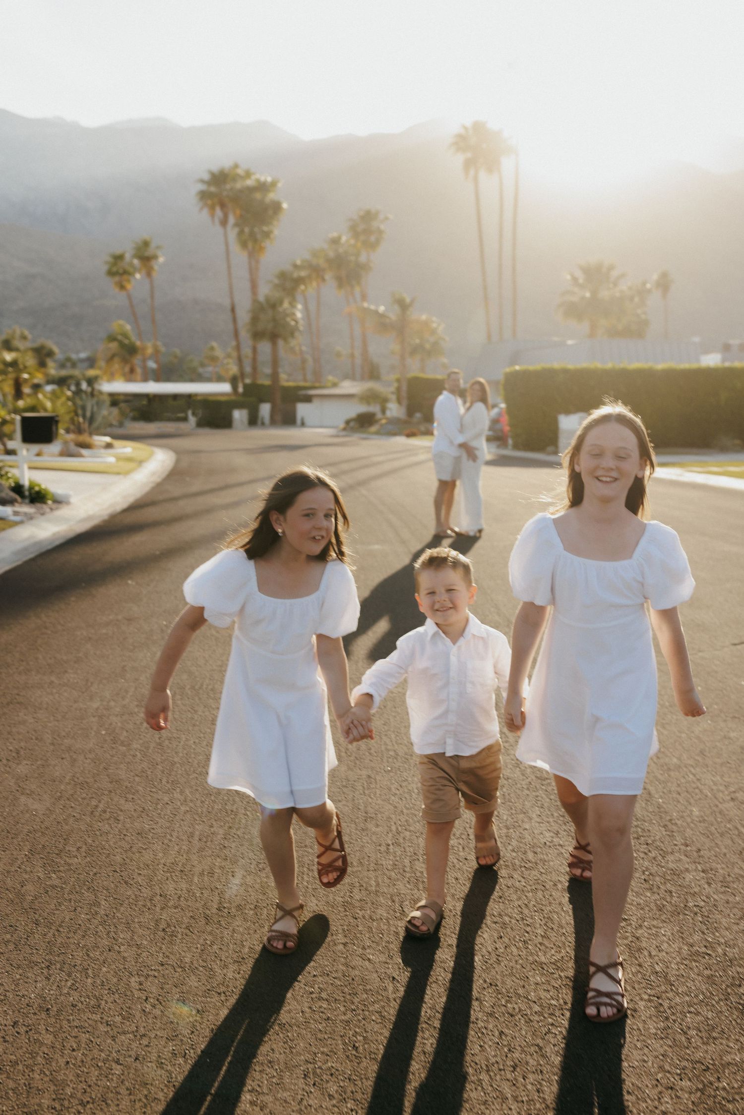 Two girls in white dresses and a boy walk hand-in-hand on a sunny Palm Springs street with mountains and palms behind.
