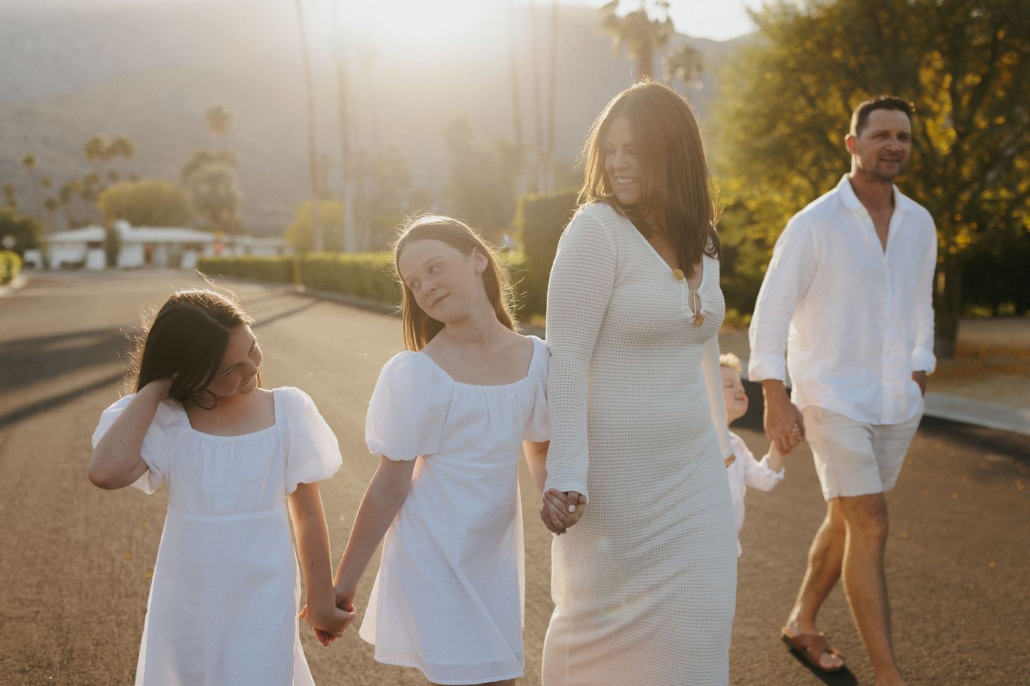 Family of four in white outfits walking hand-in-hand on a sunlit street with palm trees in the background.