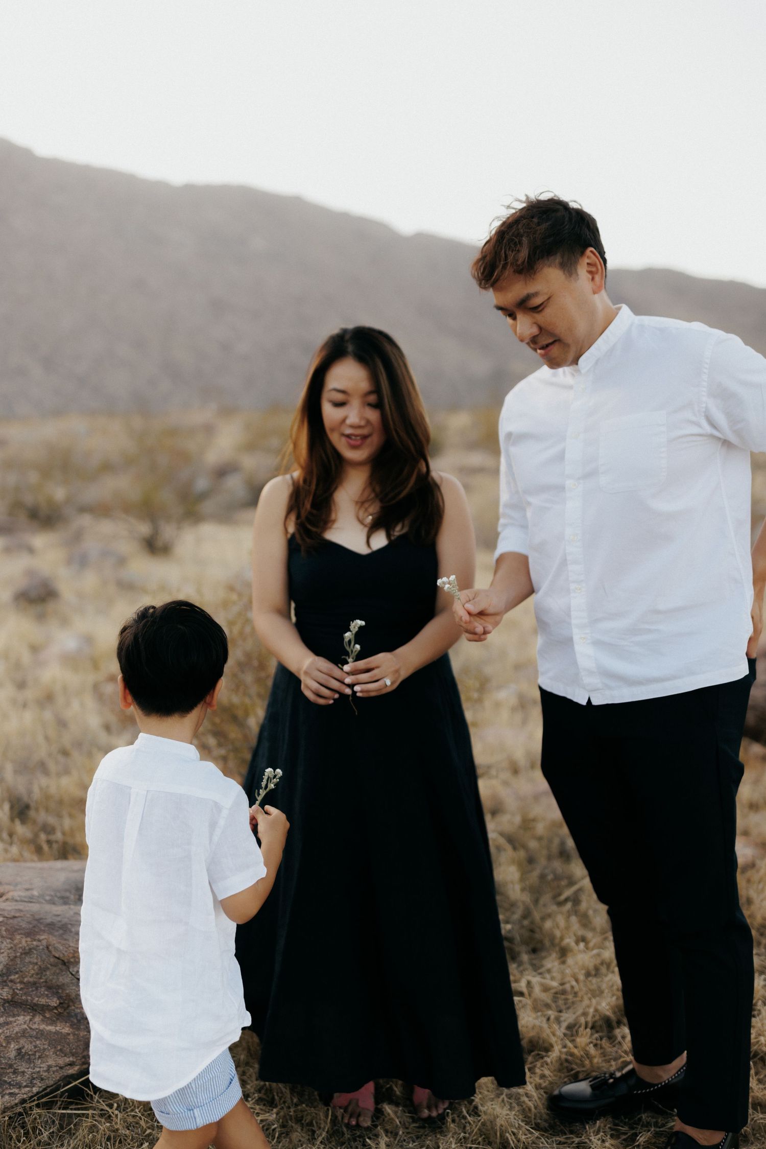 Family of three in desert setting, parents in black and white outfits look down at young boy holding wildflowers.