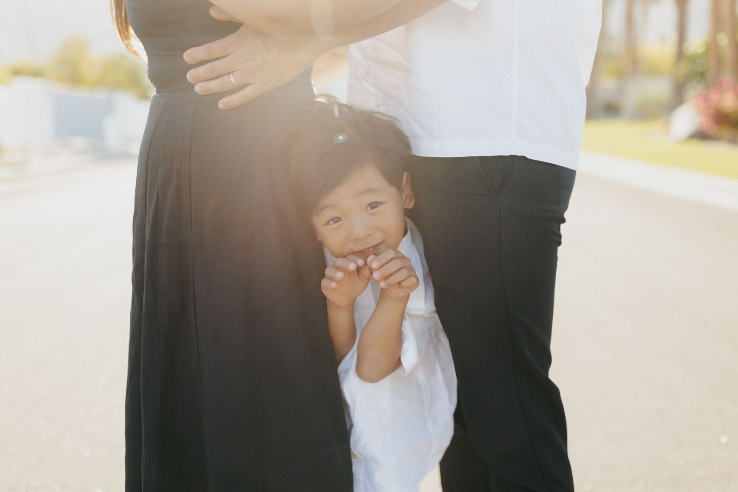Young child peeking between parents legs in warm sunlight during a family maternity photo session outdoors.