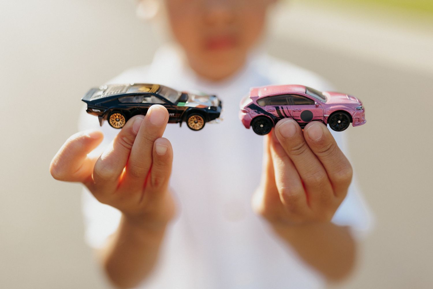 Child holding two toy cars, a black and a pink model, in outstretched hands against a blurred background.