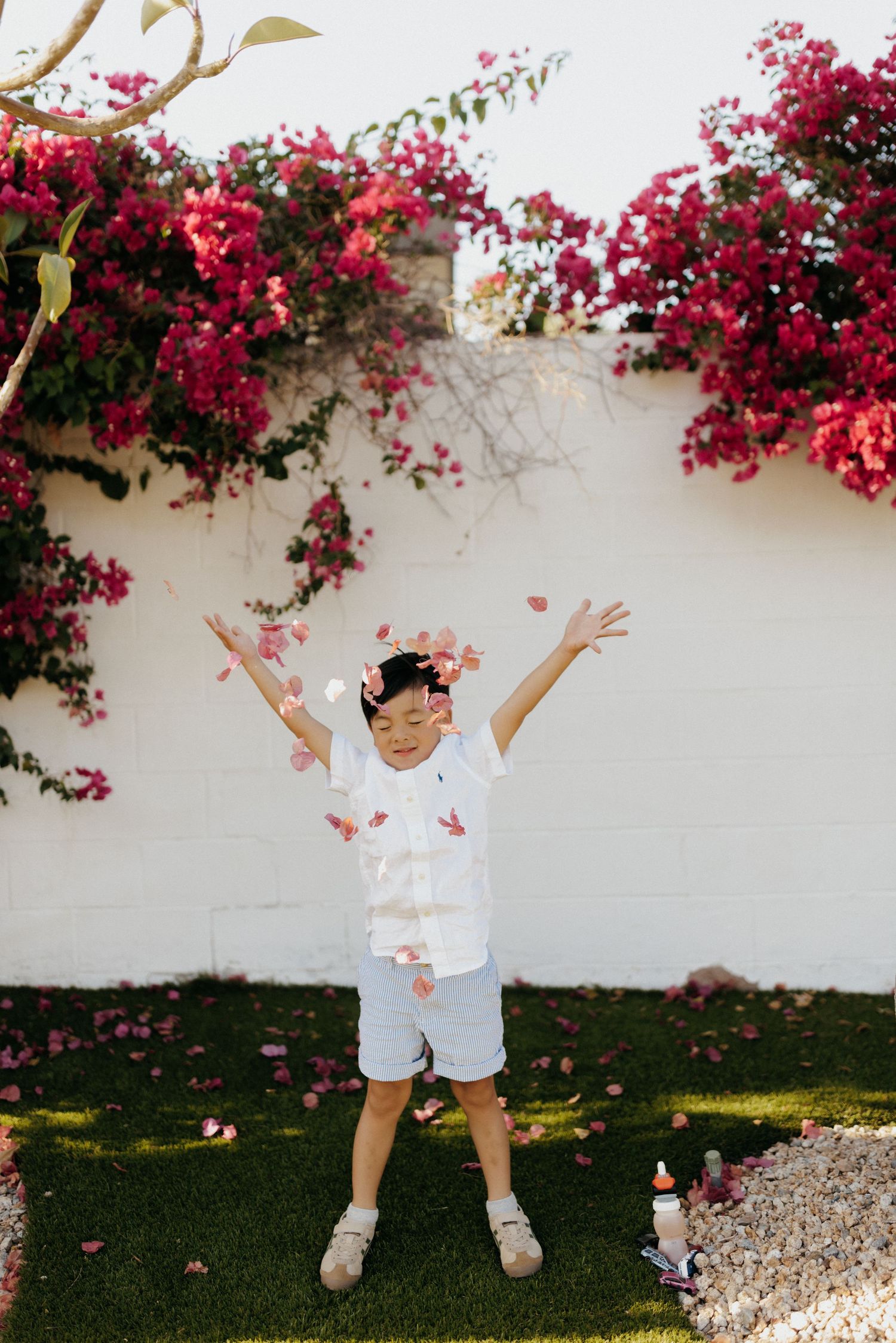 A joyful young boy in white shirt and blue shorts tosses pink flower petals in the air outdoors.