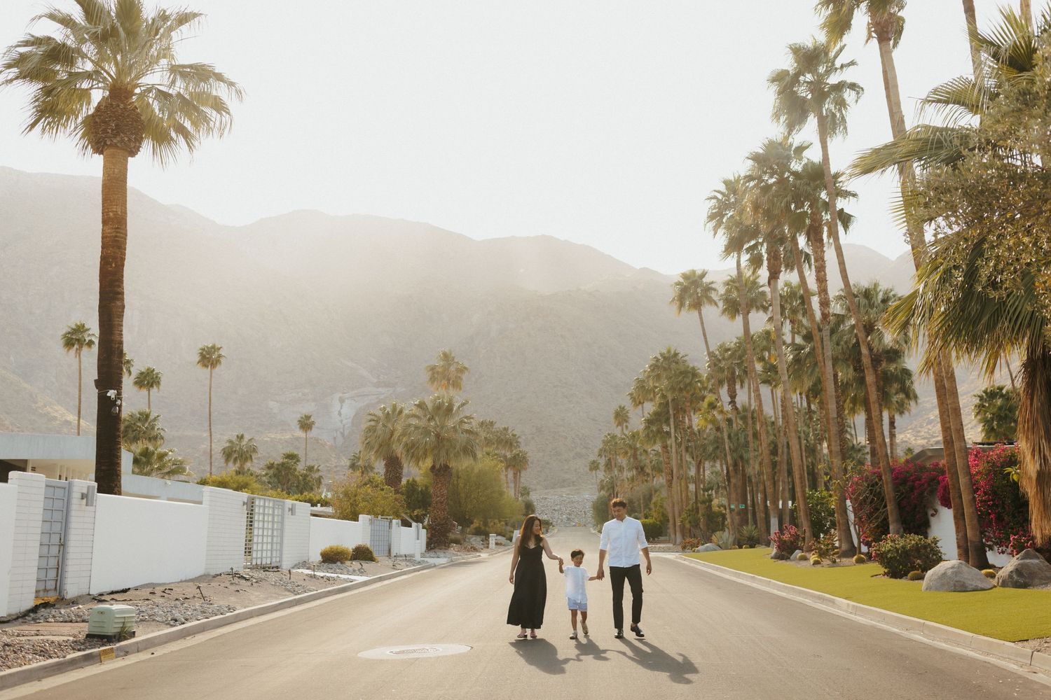 Family of three walking hand-in-hand down a palm-lined street with mountain backdrop in Palm Springs, CA.