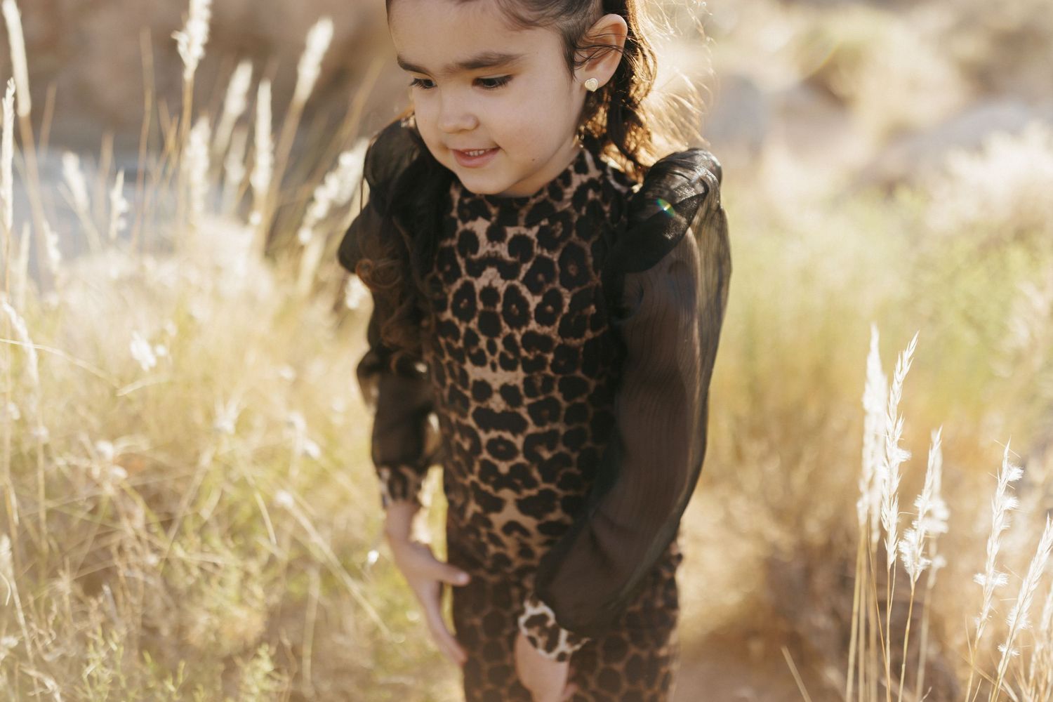 A young girl in a leopard print outfit walks through golden tall grass in a sun-drenched outdoor setting.