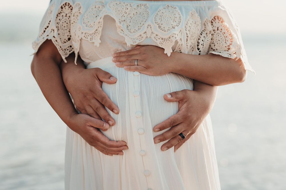 A person in a white lace dress cradles their pregnant belly while standing near water at sunset.