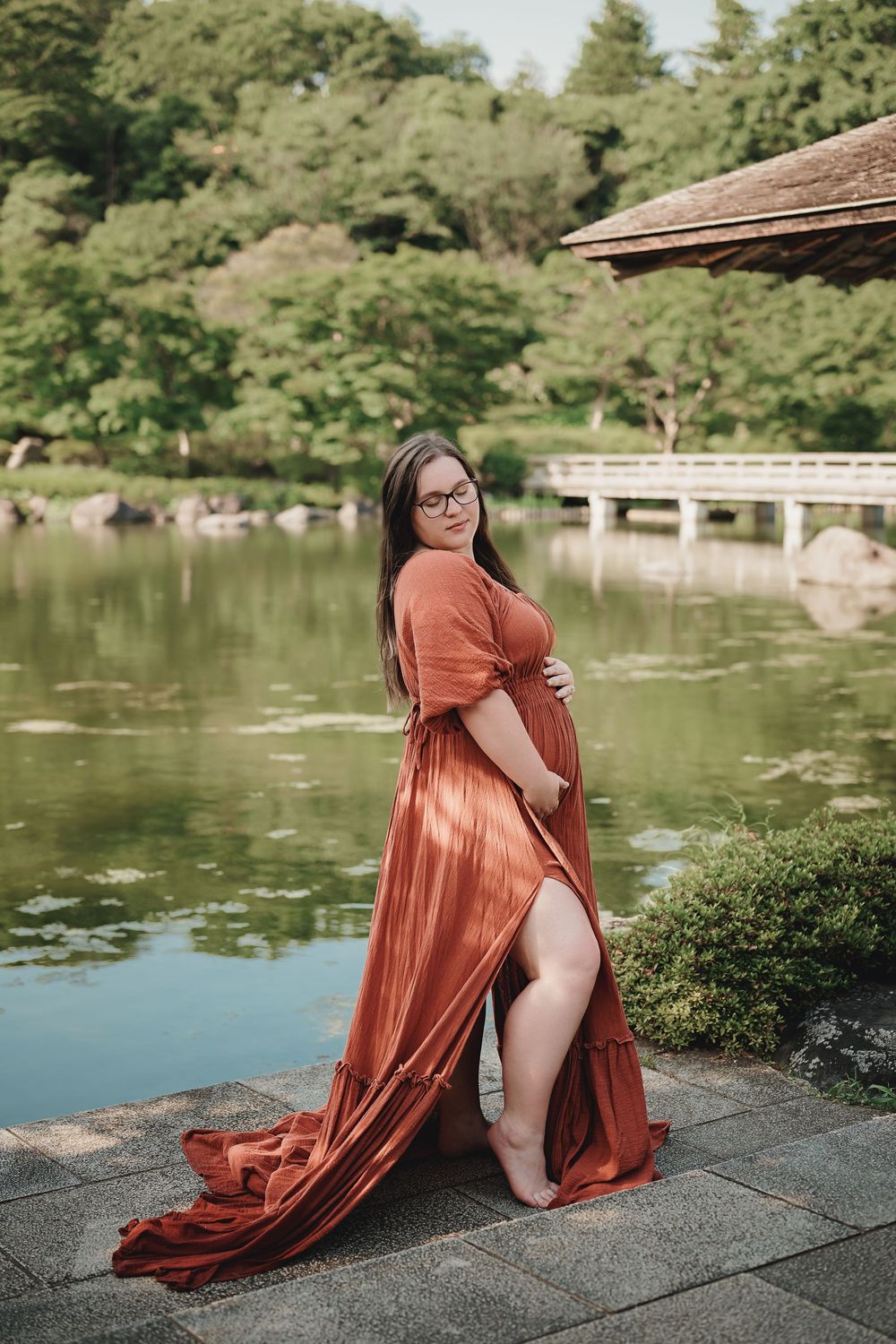 A pregnant woman in a flowing coral dress poses elegantly by a serene Japanese garden pond with bridge in background.