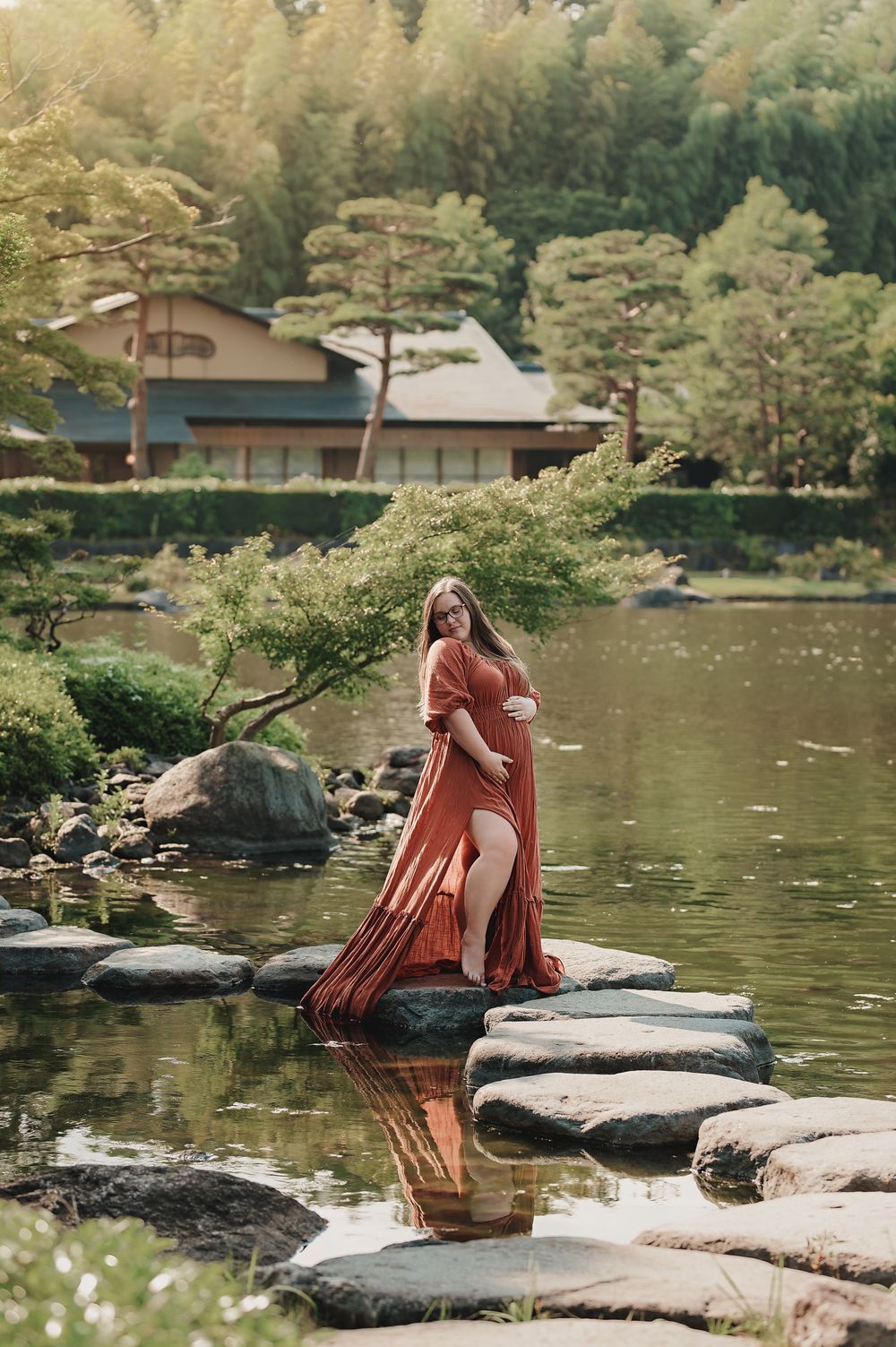 Someone in a coral dress sits contemplatively on large rocks by a serene lake surrounded by pine trees.