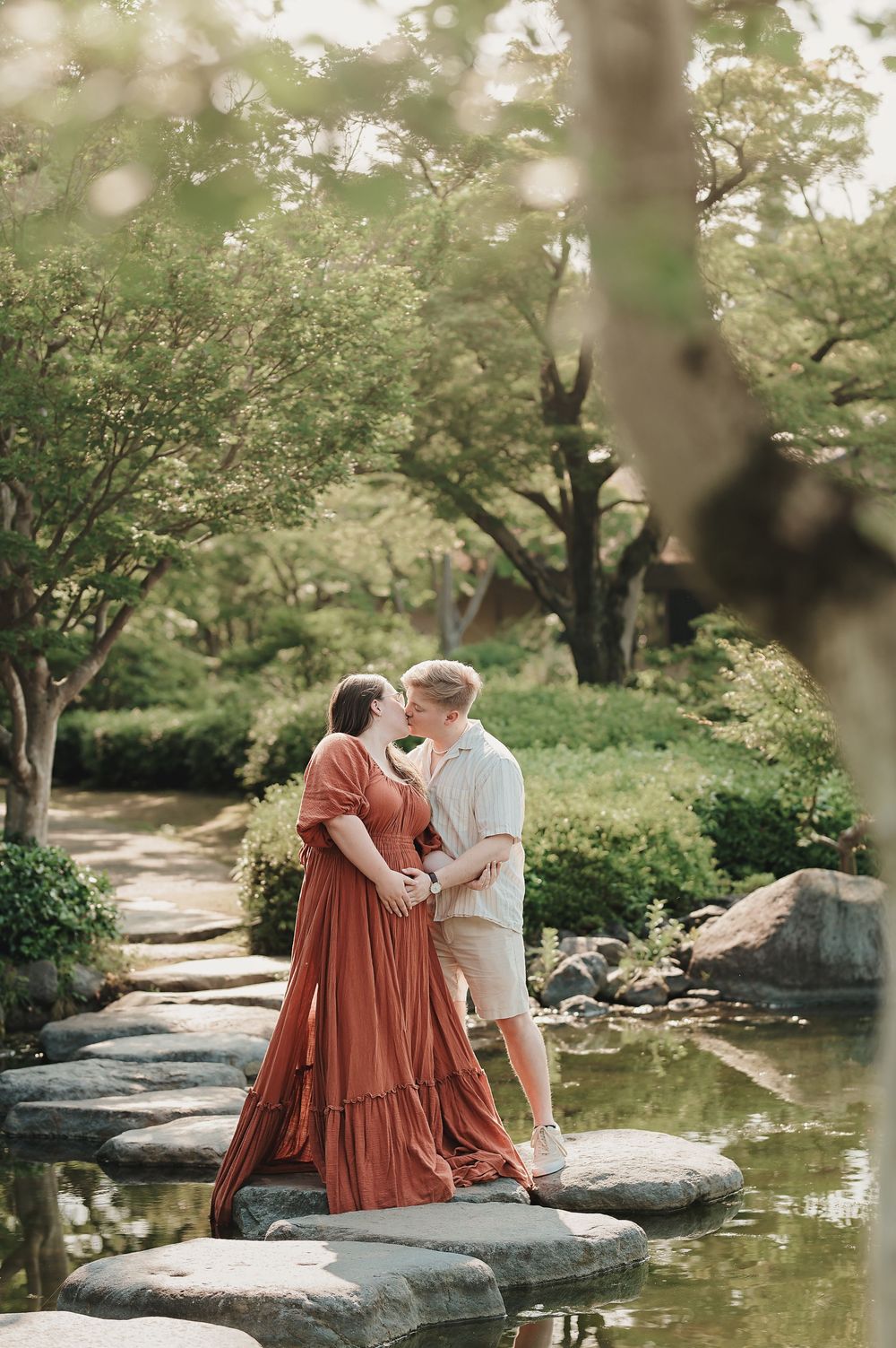 A couple shares a tender moment while standing on rocks beside a tranquil pond surrounded by lush greenery.
