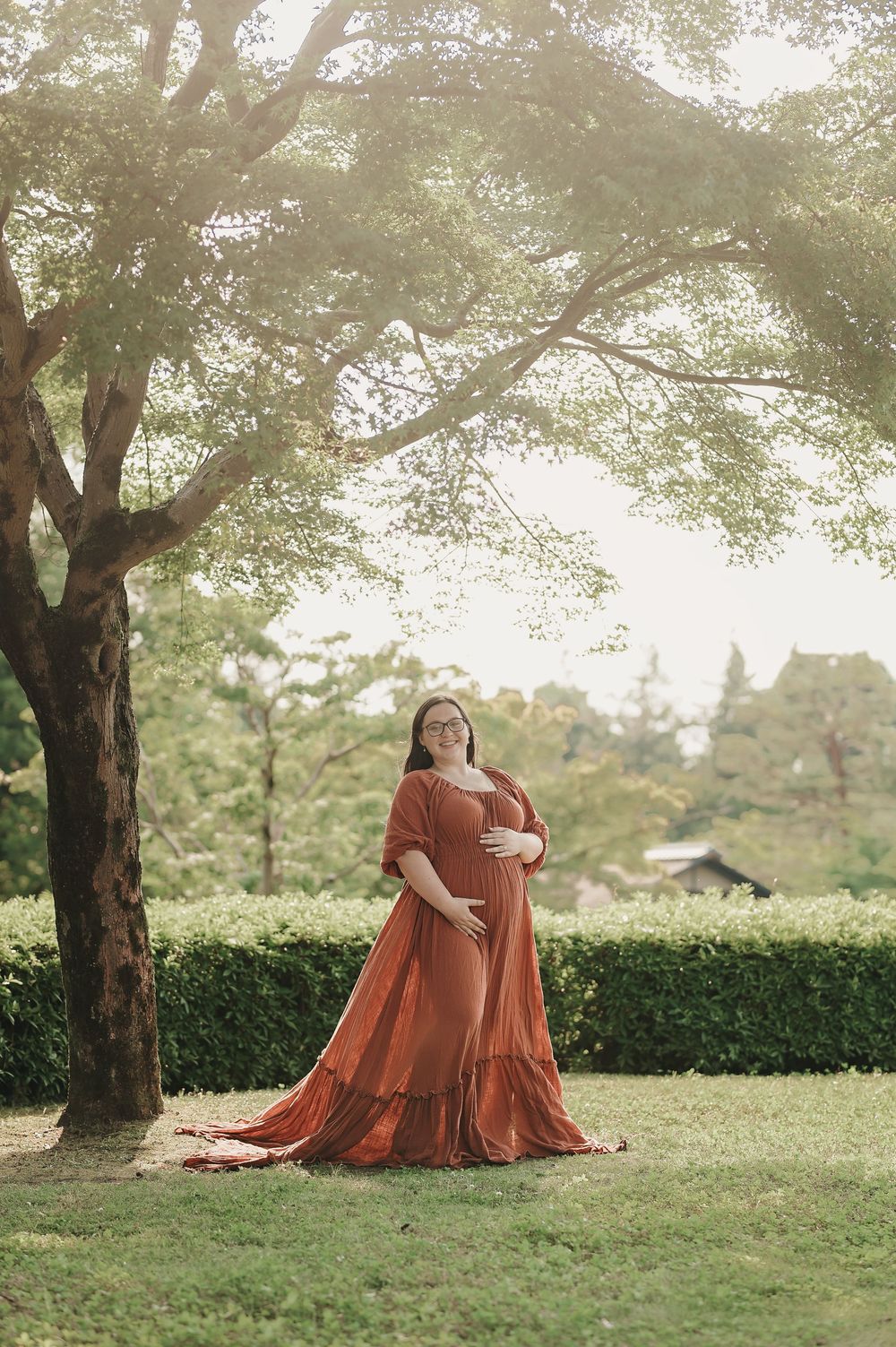 A pregnant figure in a flowing rust-colored dress poses beneath a large tree in a sunlit garden setting.