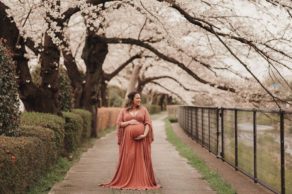 A person in a flowing coral dress stands on a path beneath blooming cherry blossom trees in spring.