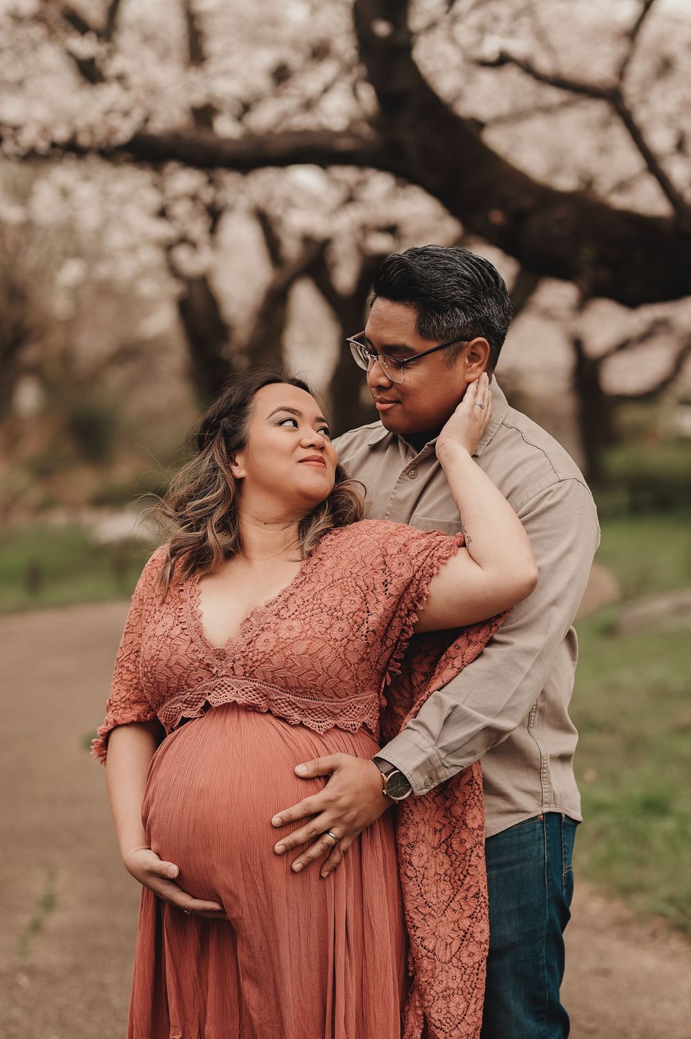 A couple shares an intimate moment outdoors during a maternity photoshoot, with cherry blossom trees in the background.