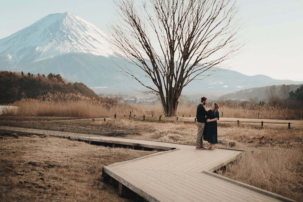 Two silhouettes stand on a wooden boardwalk with Mount Fuji and a bare tree rising majestically in the background.